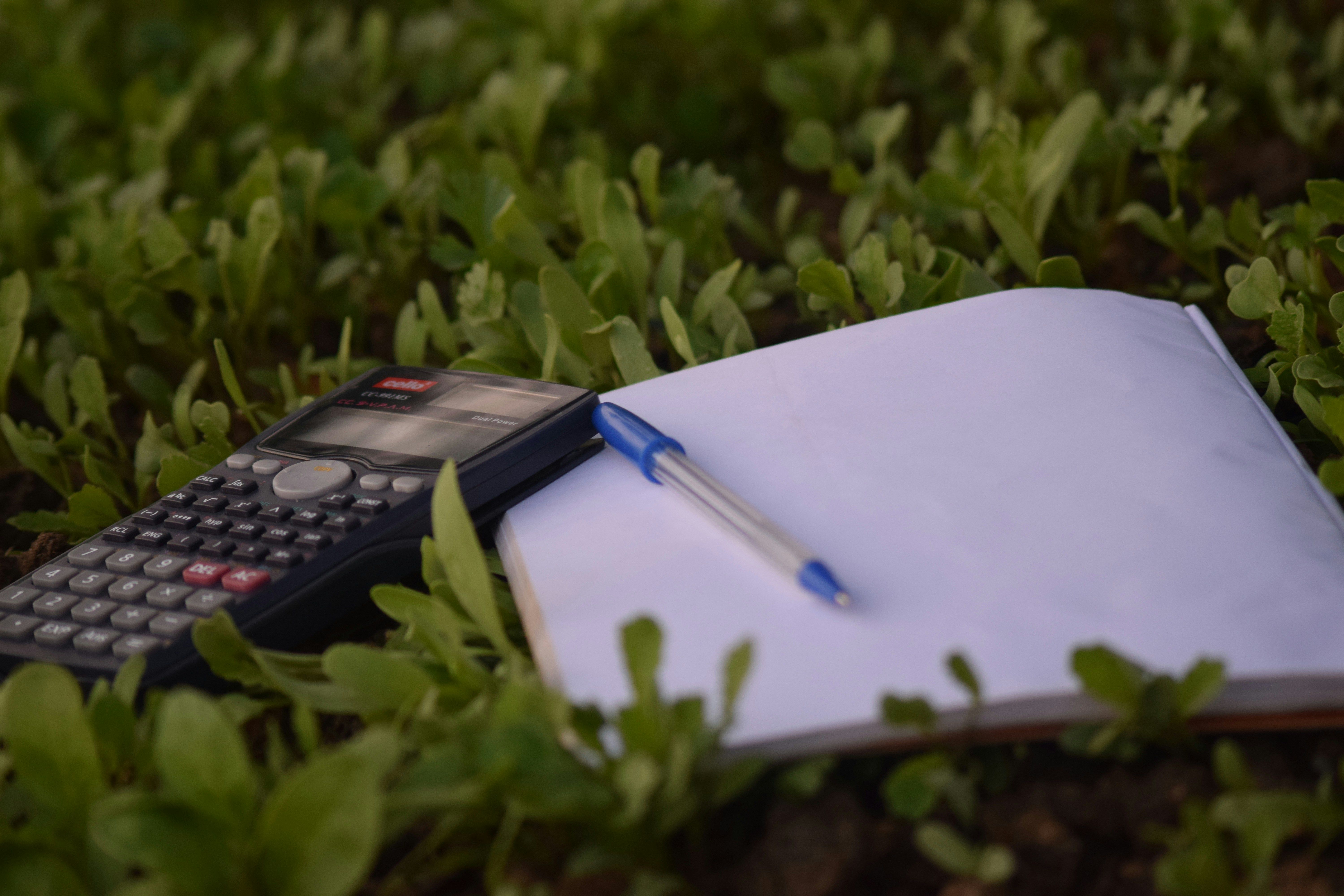Calculator with notebook and pen laying on a field of grass.