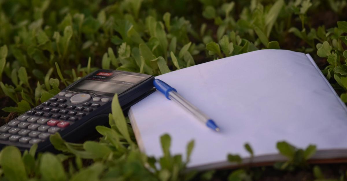 Calculator with notebook and pen laying on a field of grass.