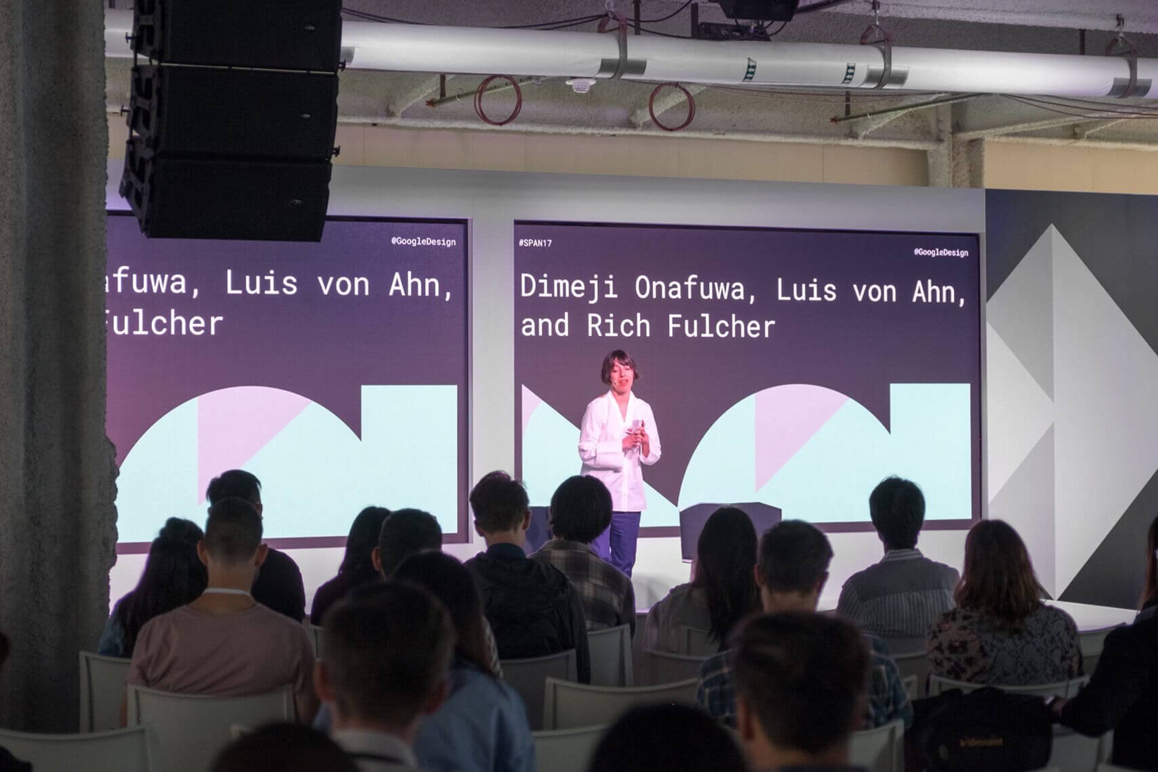 Photo of stage with woman giving speech and Google Span shapes on displays