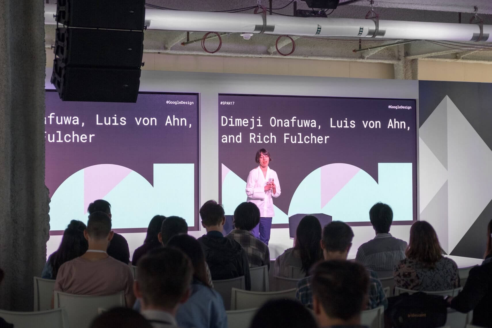 Photo of stage with woman giving speech and Google Span shapes on displays
