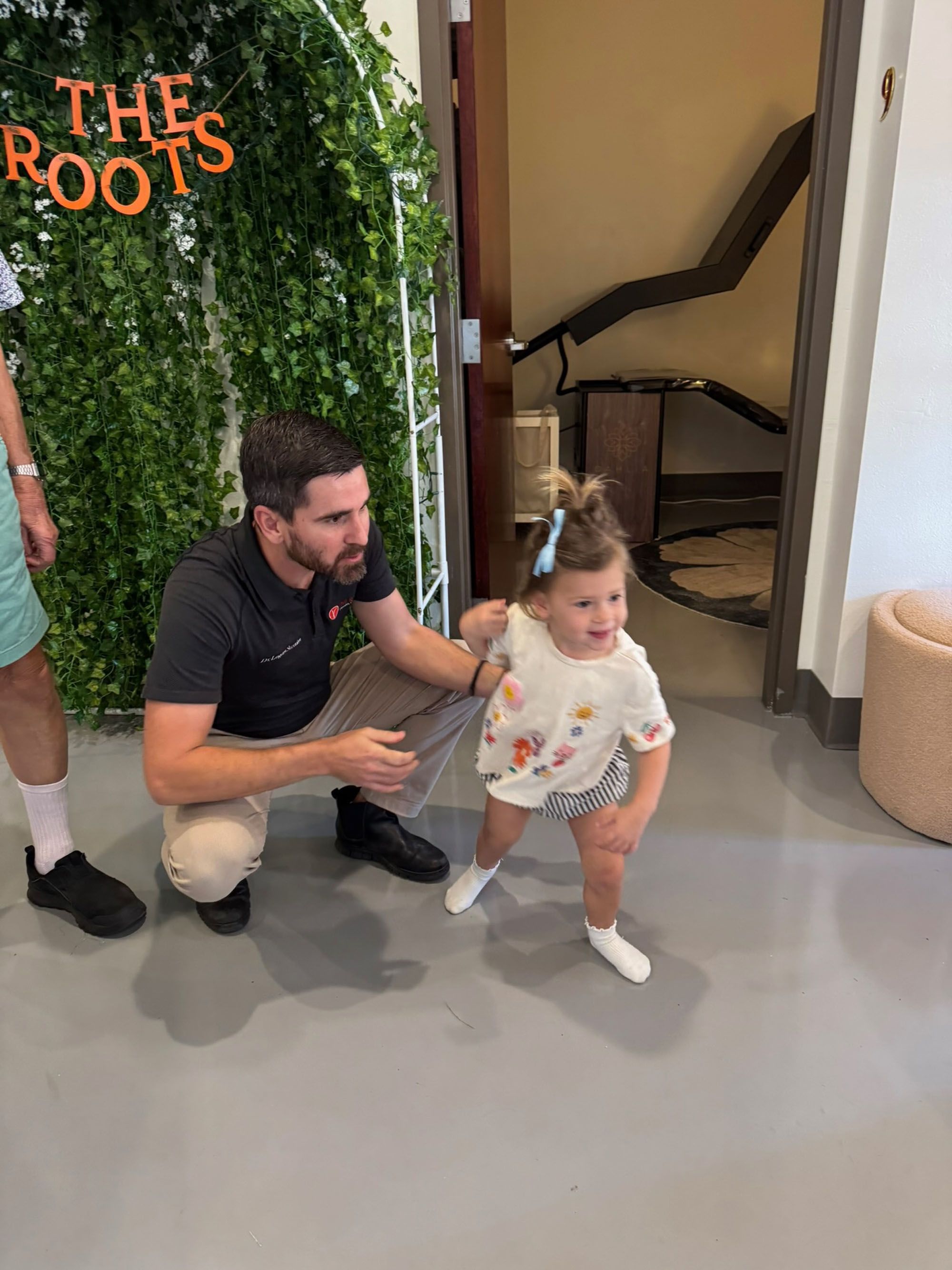 Dr. Fox kneels by The Roots arch as a toddler runs past at a family event.