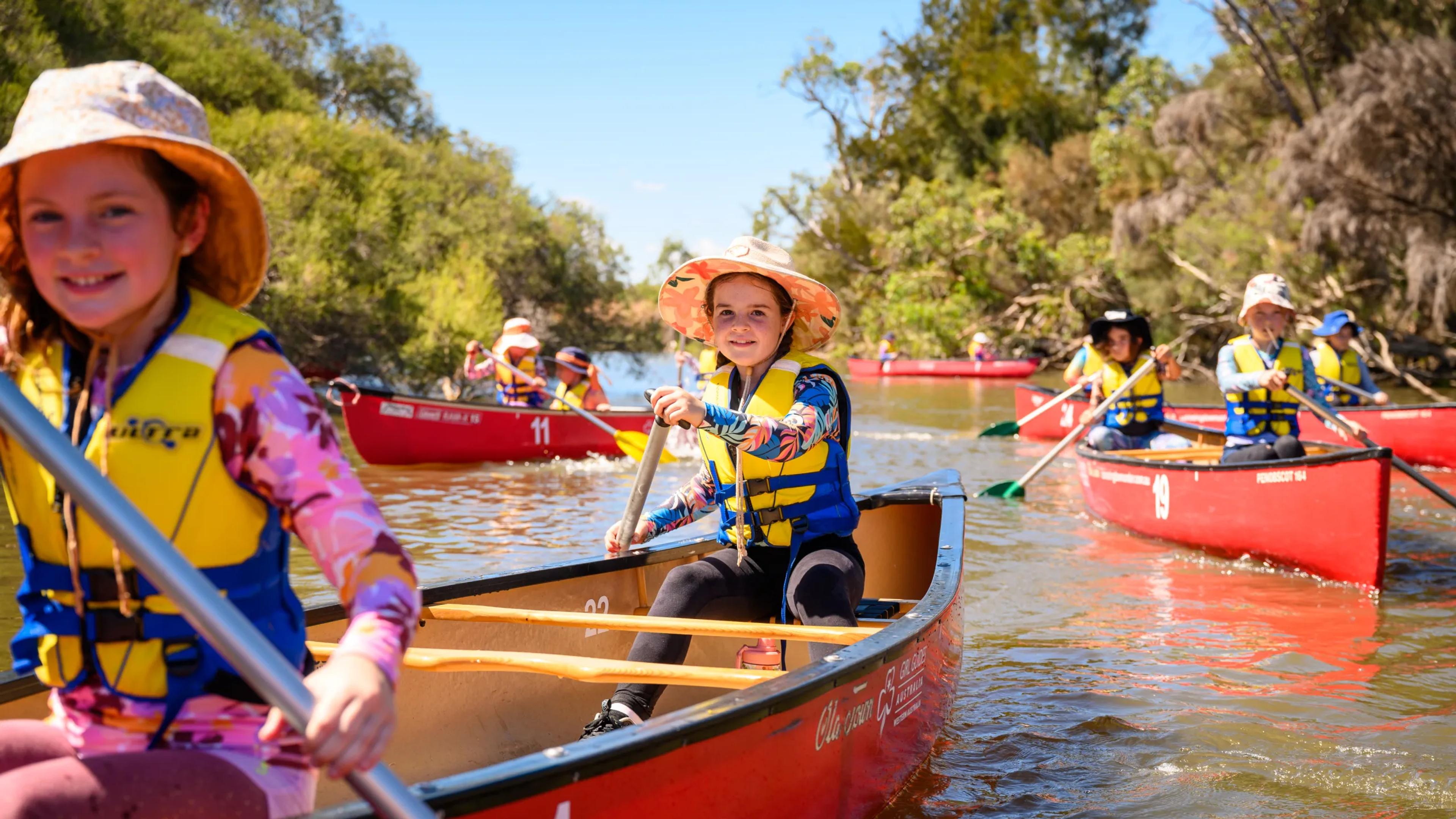 A photo of a series of canoes, with two girls in each canoe, rowing down a river.