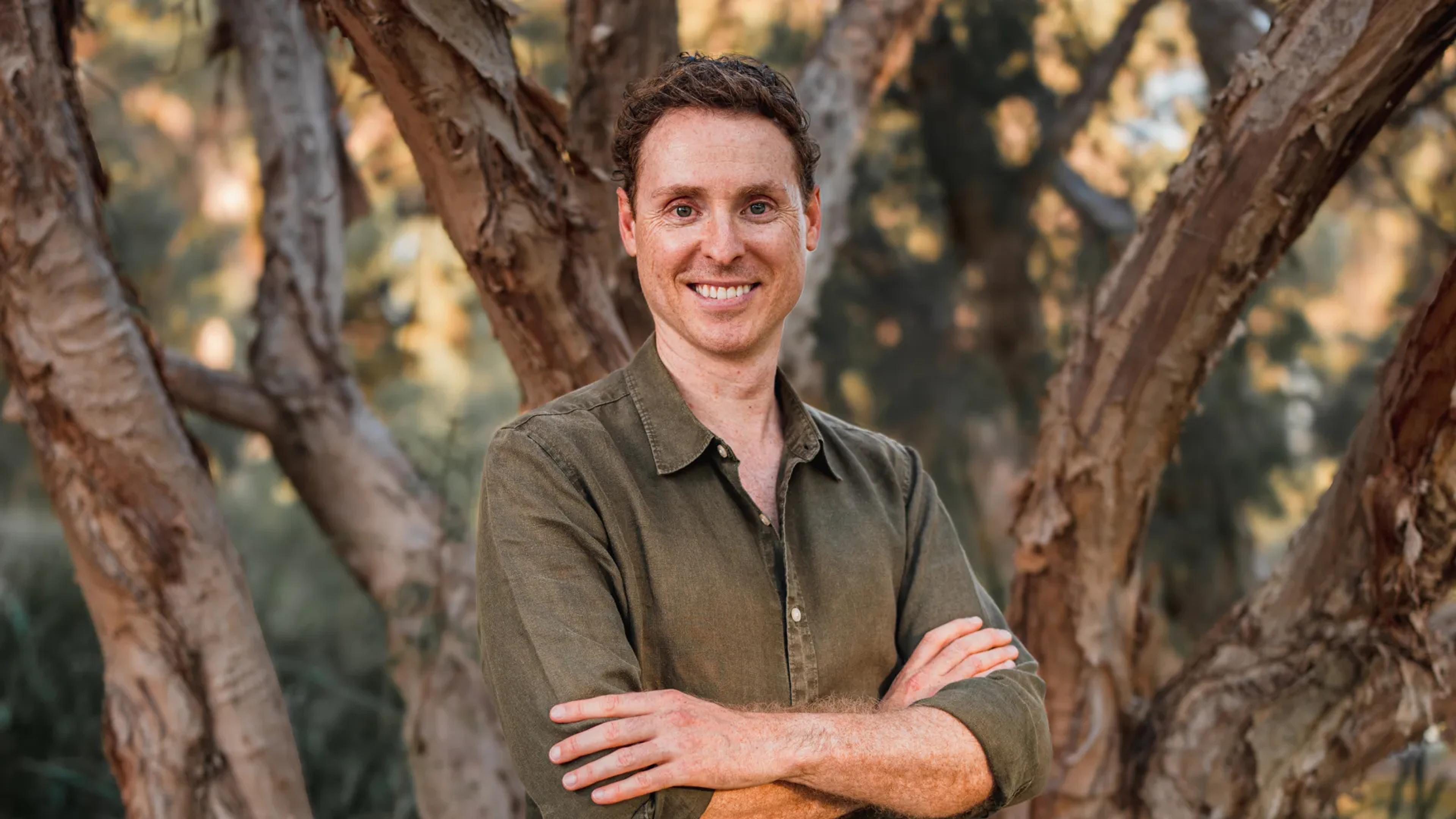A photo of a man standing in the bush with his arms crossed.