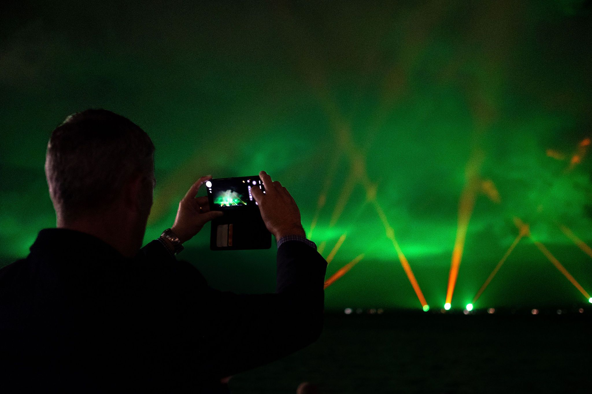 A man at night holds up his camera phone with red and green lights in the background