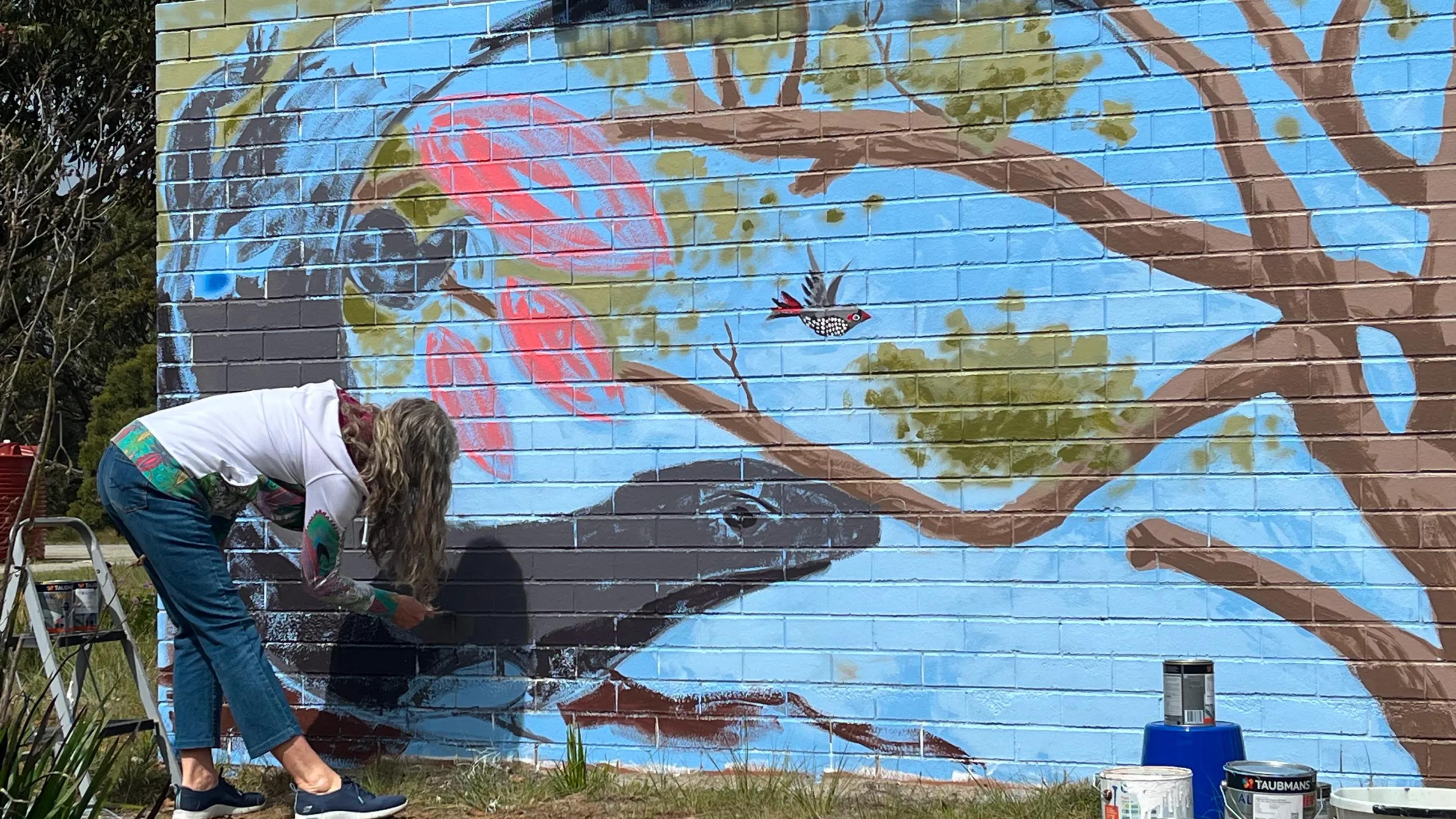 A photo of a woman bending over, painting a mural on a brick wall.
