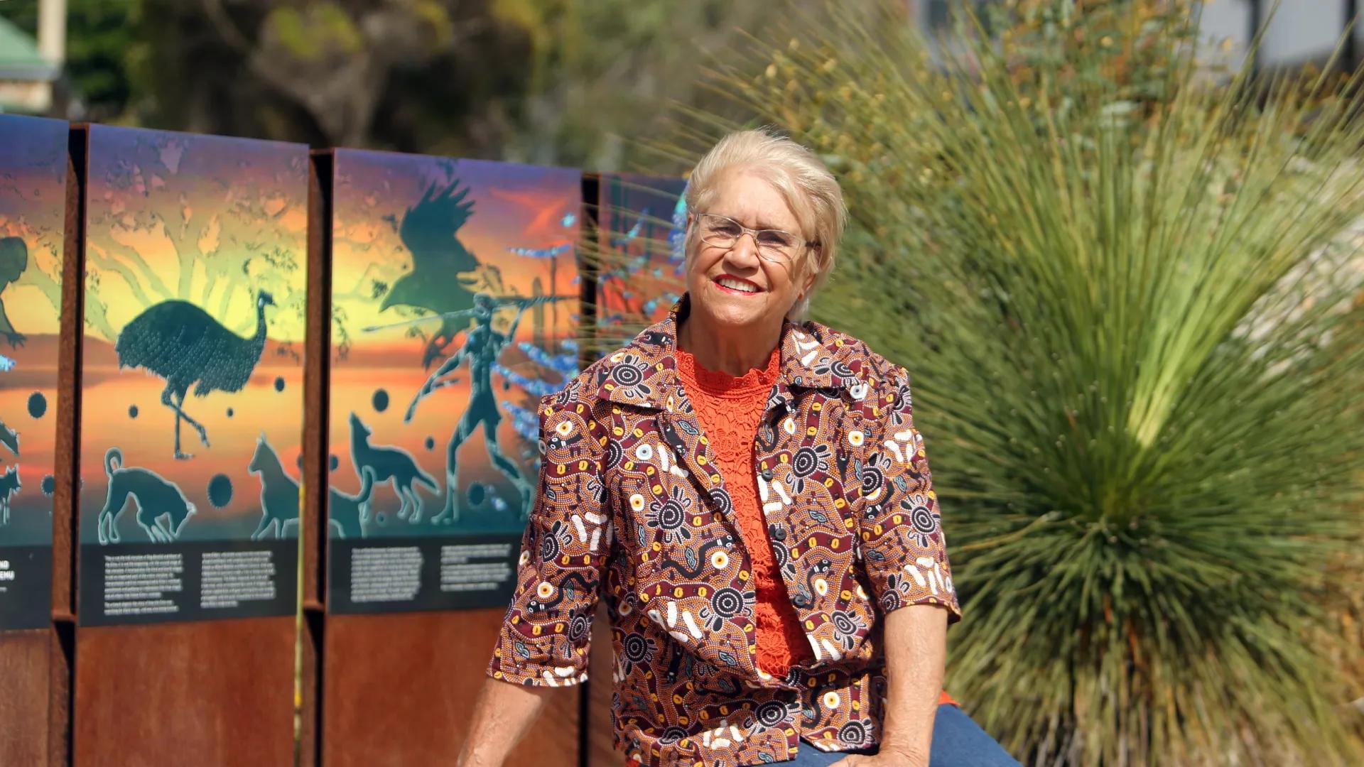 A photo of an elderly woman standing in front of some bush and indigenous artwork.