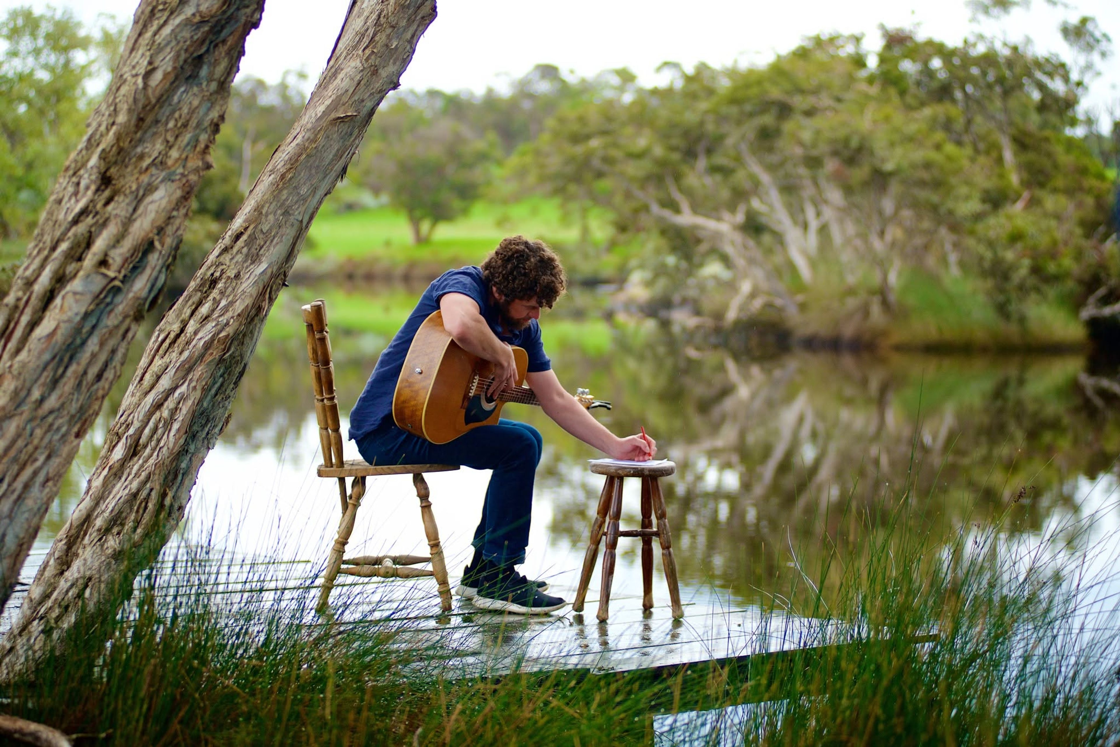 A guitarist sits by a lake writing music