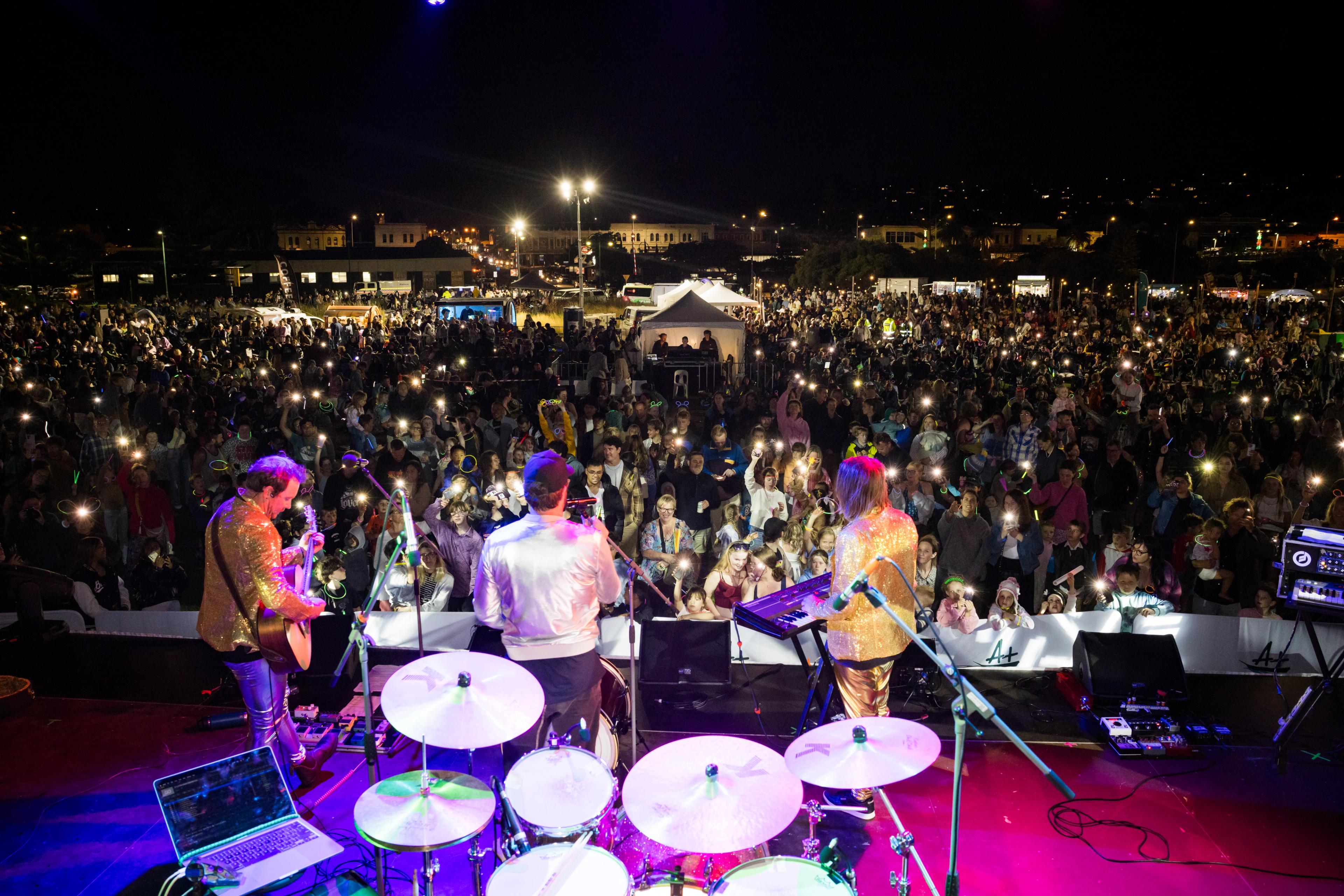 A photo taken from behind a band, with a large crowd before them, illuminated by glow sticks and torch lights. 