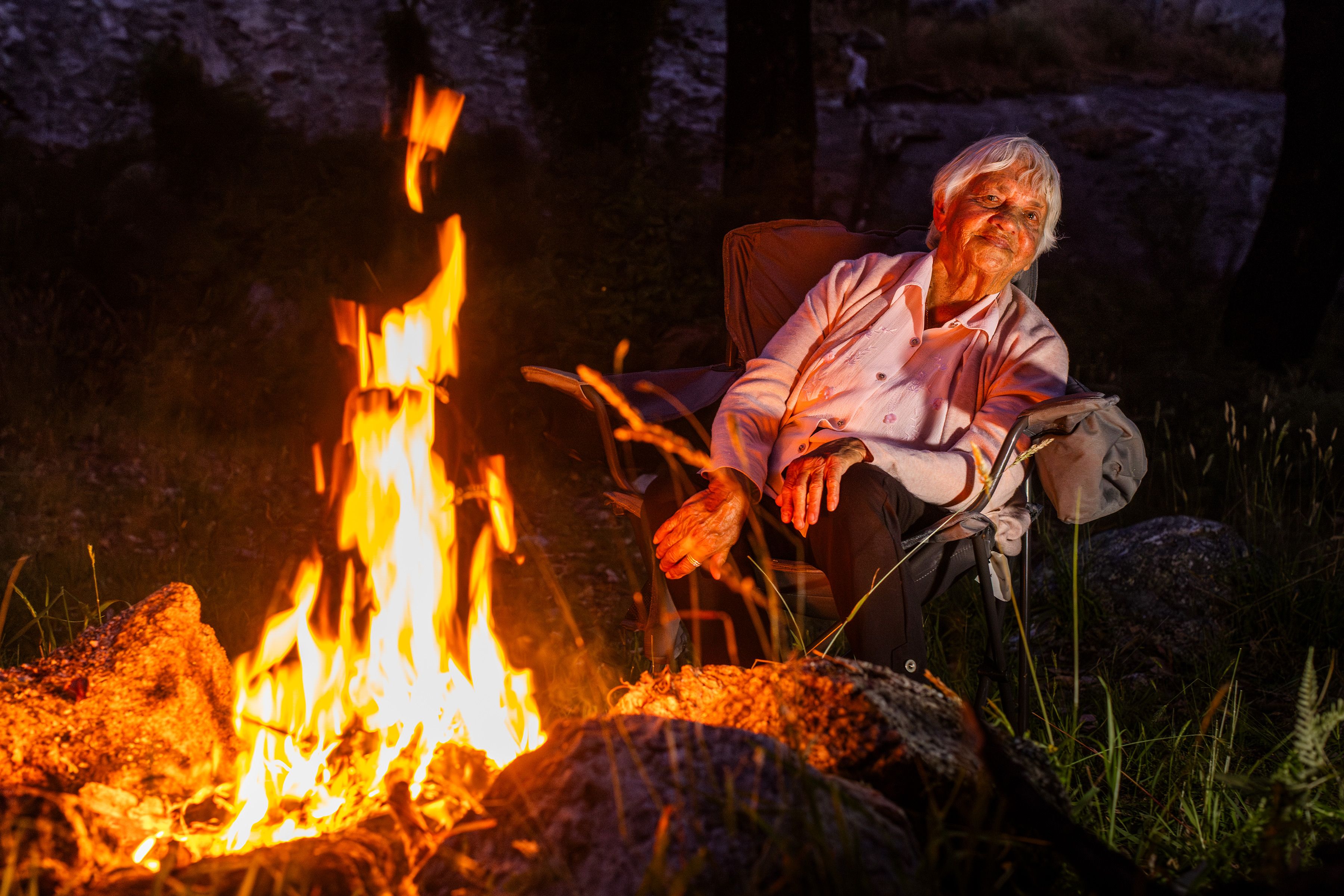 A Menang Elder illuminated by firelight, looking around an open fire, towards the camera