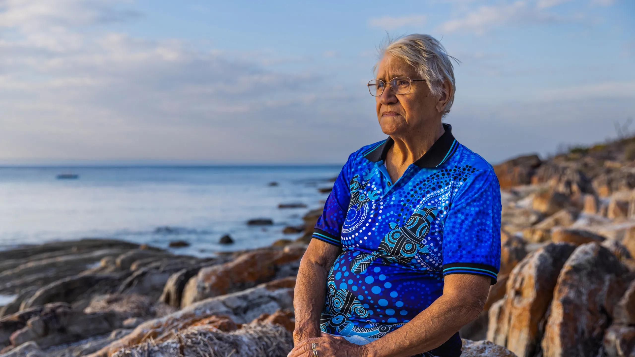 A photo of a woman sitting on a rock at the beach, looking out at the sunset.