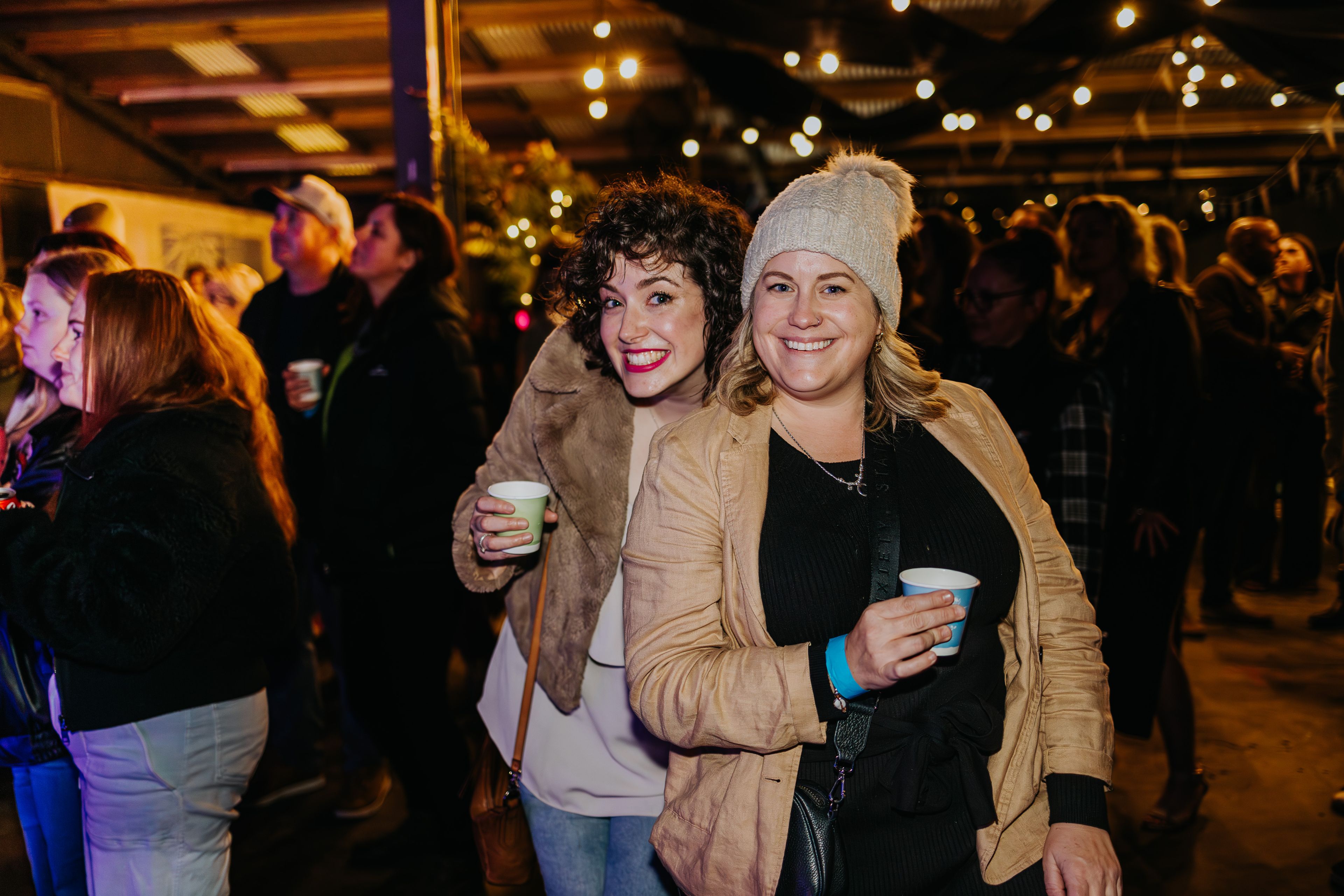 Two people in warm cardigans and beanies having a drink in a crowded venue.