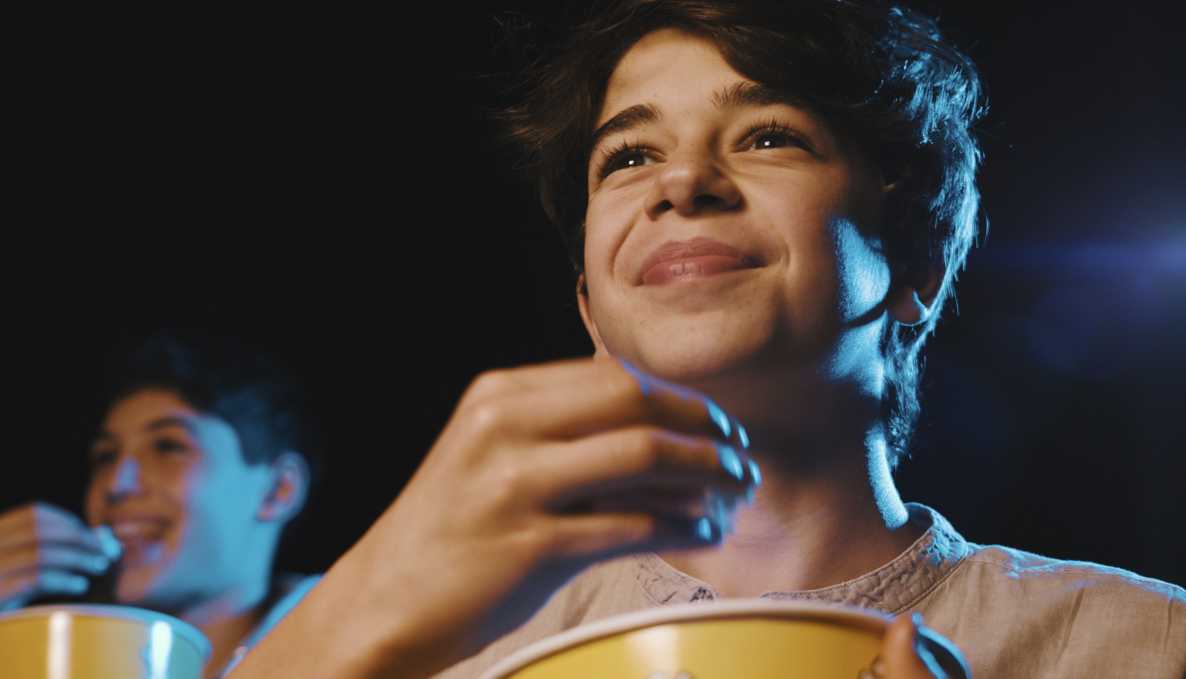Close up of a young boy, smiling as he looks towards a screen and eats popcorn.
