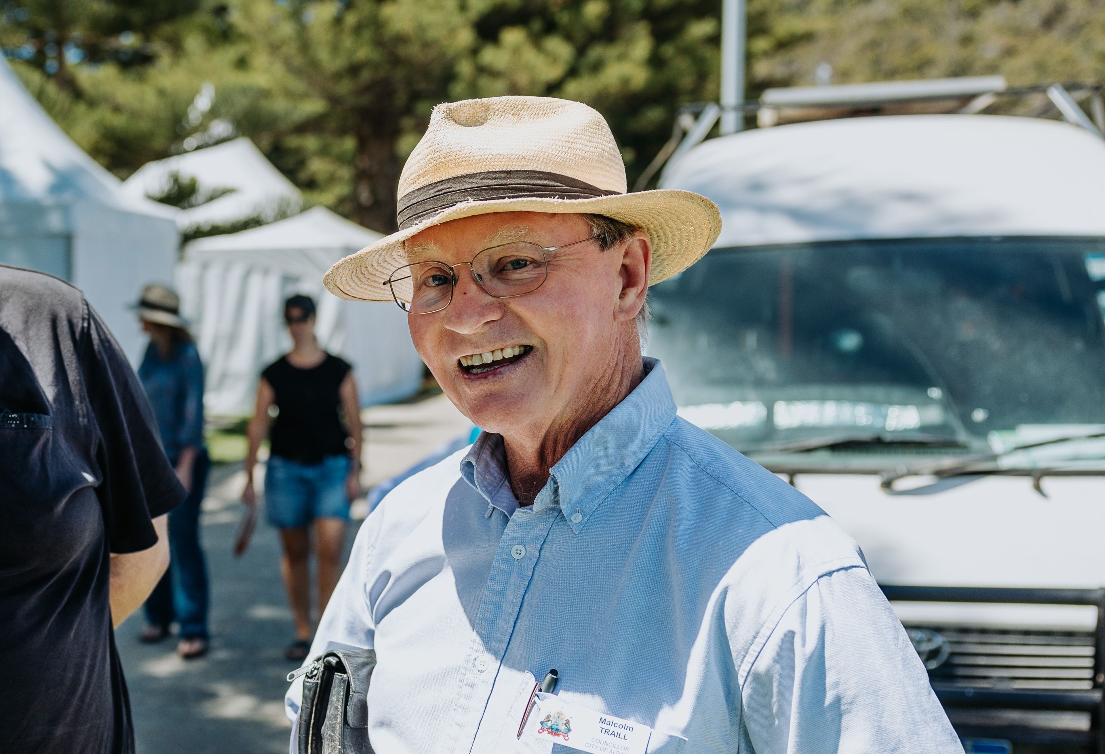 A smiling older man wearing a straw hat, glasses and a light blue shirt holds a takeaway coffee cup while standing outdoors at a sunny event.