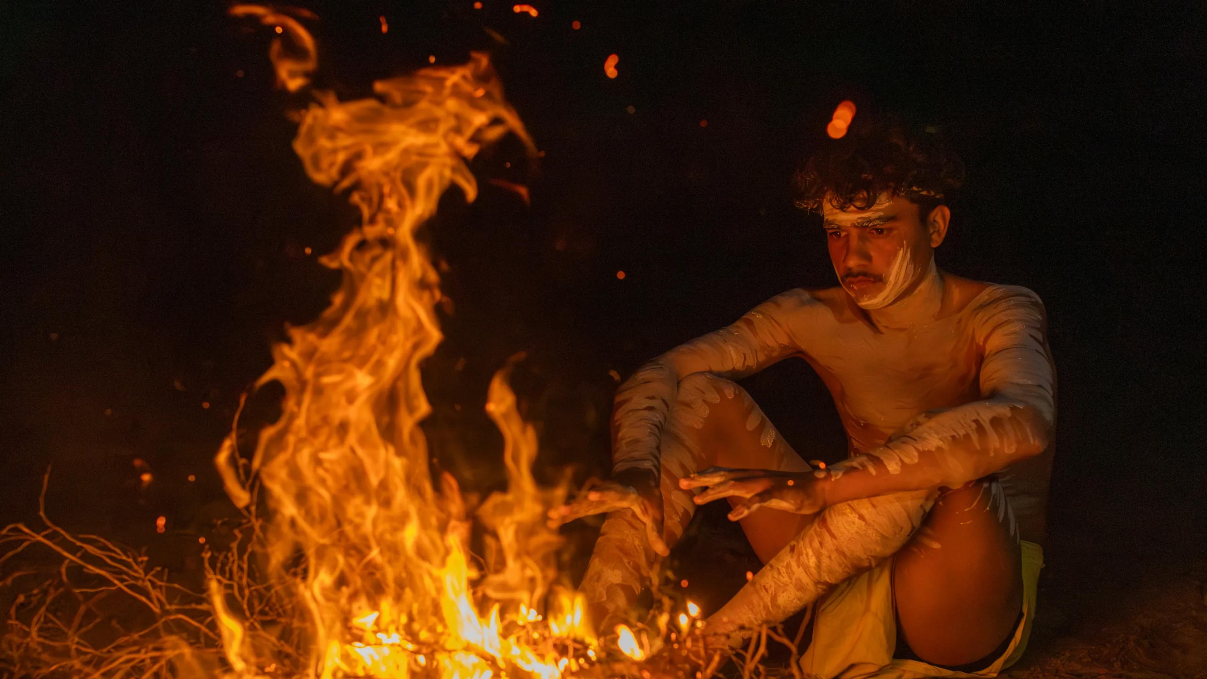 A photo of an indigenous man sitting in front of a fire warming his hands.