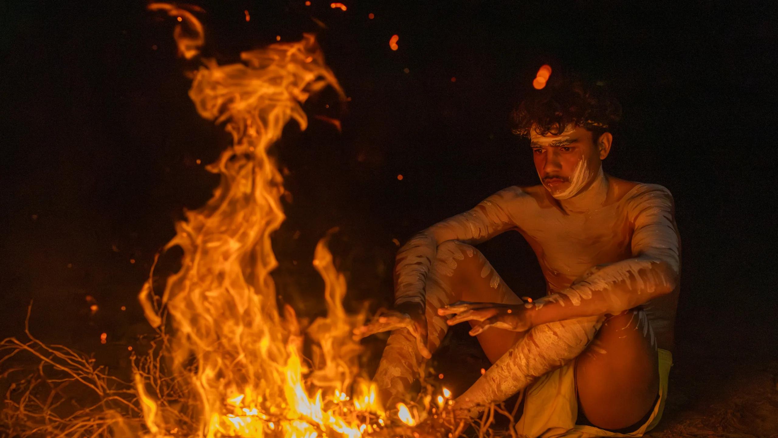 A photo of an indigenous man sitting in front of a fire warming his hands.
