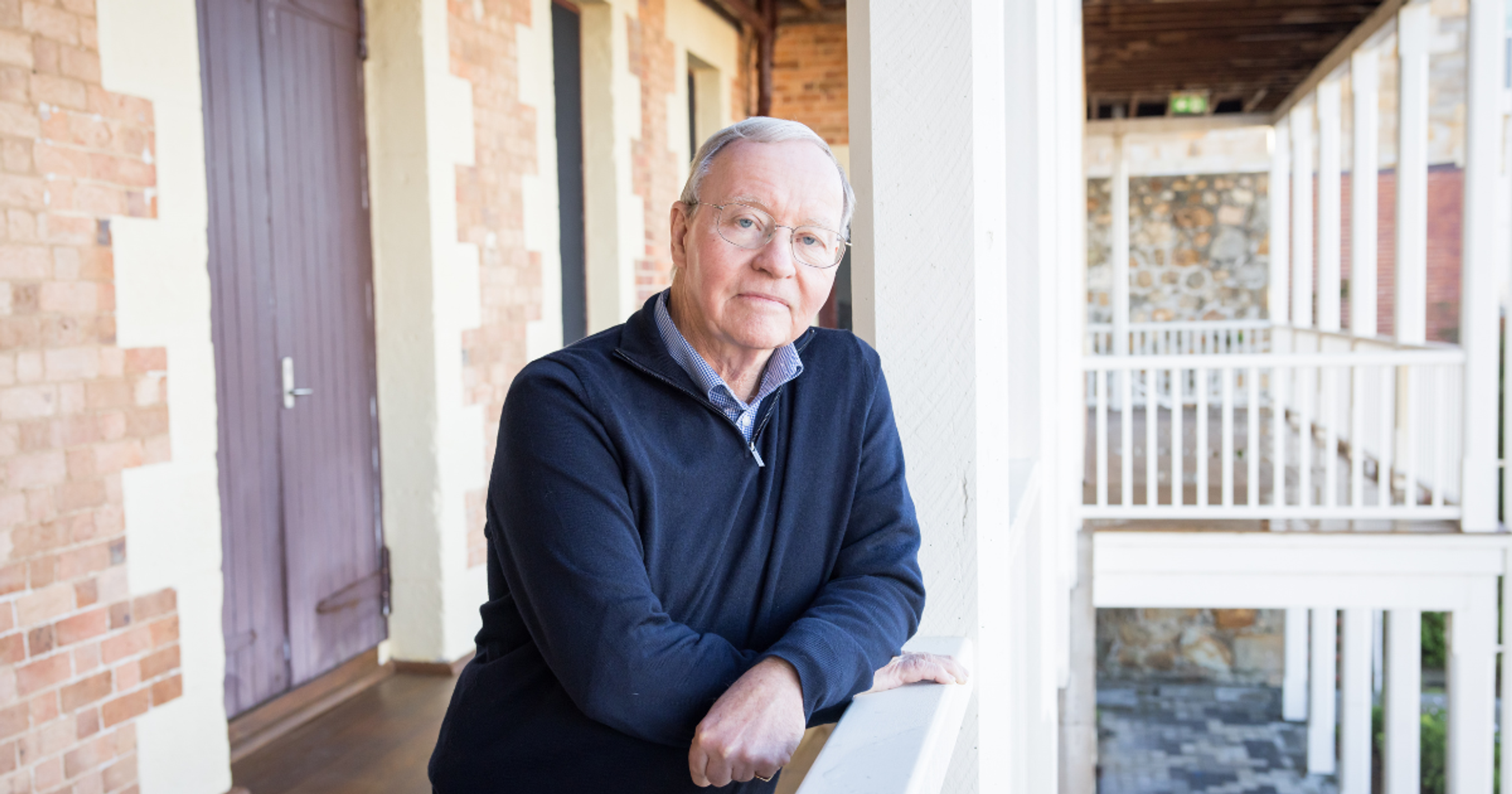Older man wearing glasses and leaning on a railing in front of a stone building
