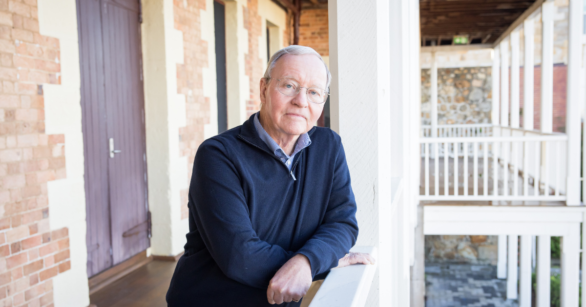 Older man wearing glasses and leaning on a railing in front of a stone building