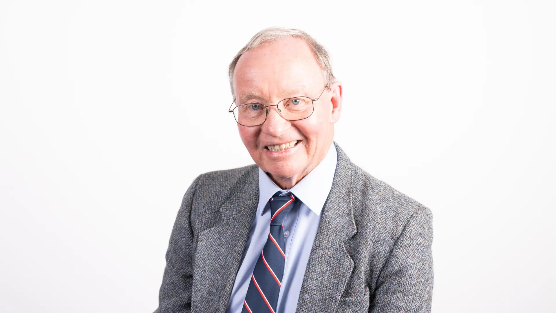 A photo of an elderly man wearing glasses sitting in front of a white background.