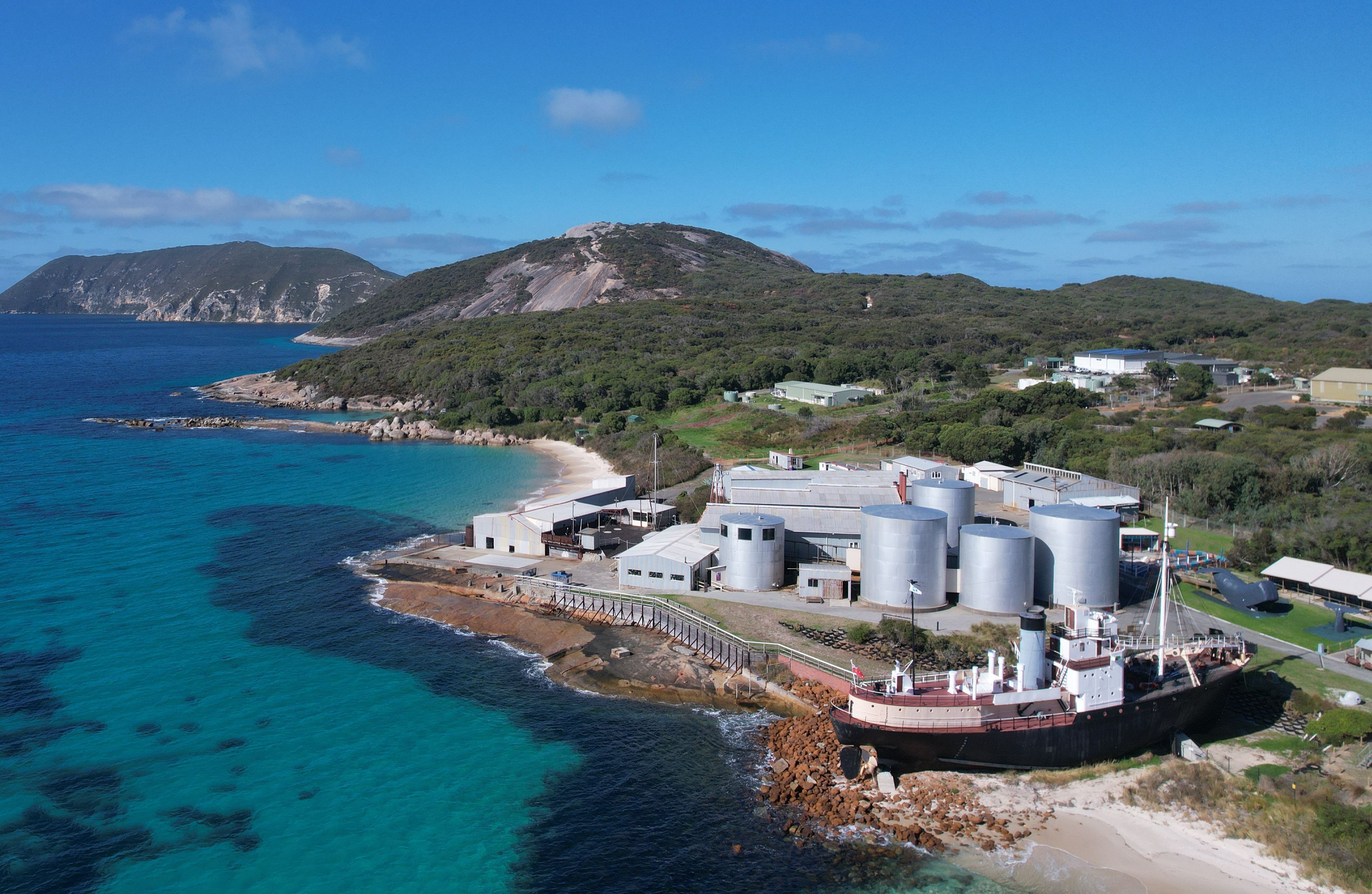Aerial view of a coastline with a large boat sitting on the land and various sheds and tanks in front of green hills.