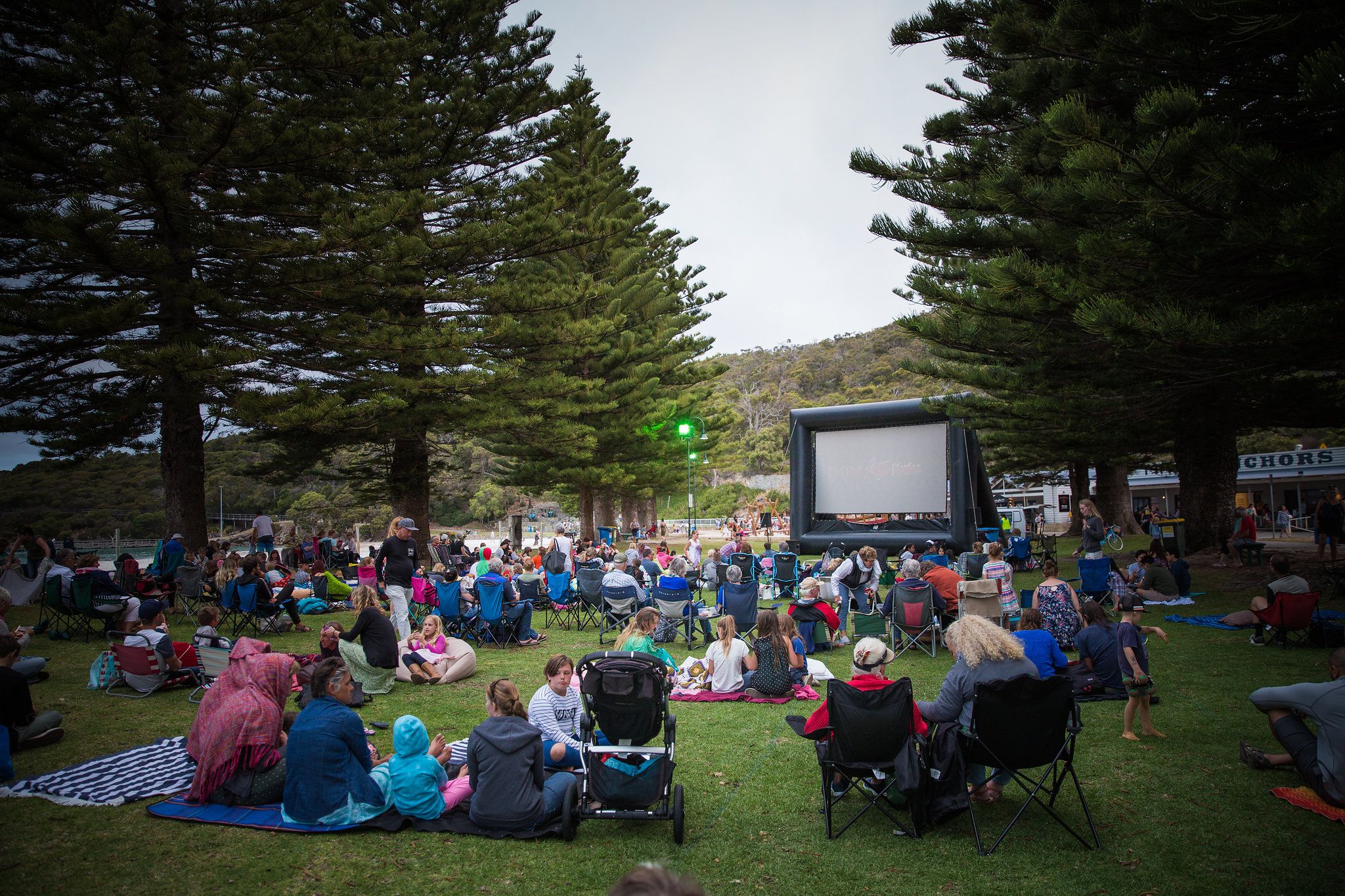 Community are gathered on picnic blankets at Binalup / Middleton Beach in front of a large outdoor screen.