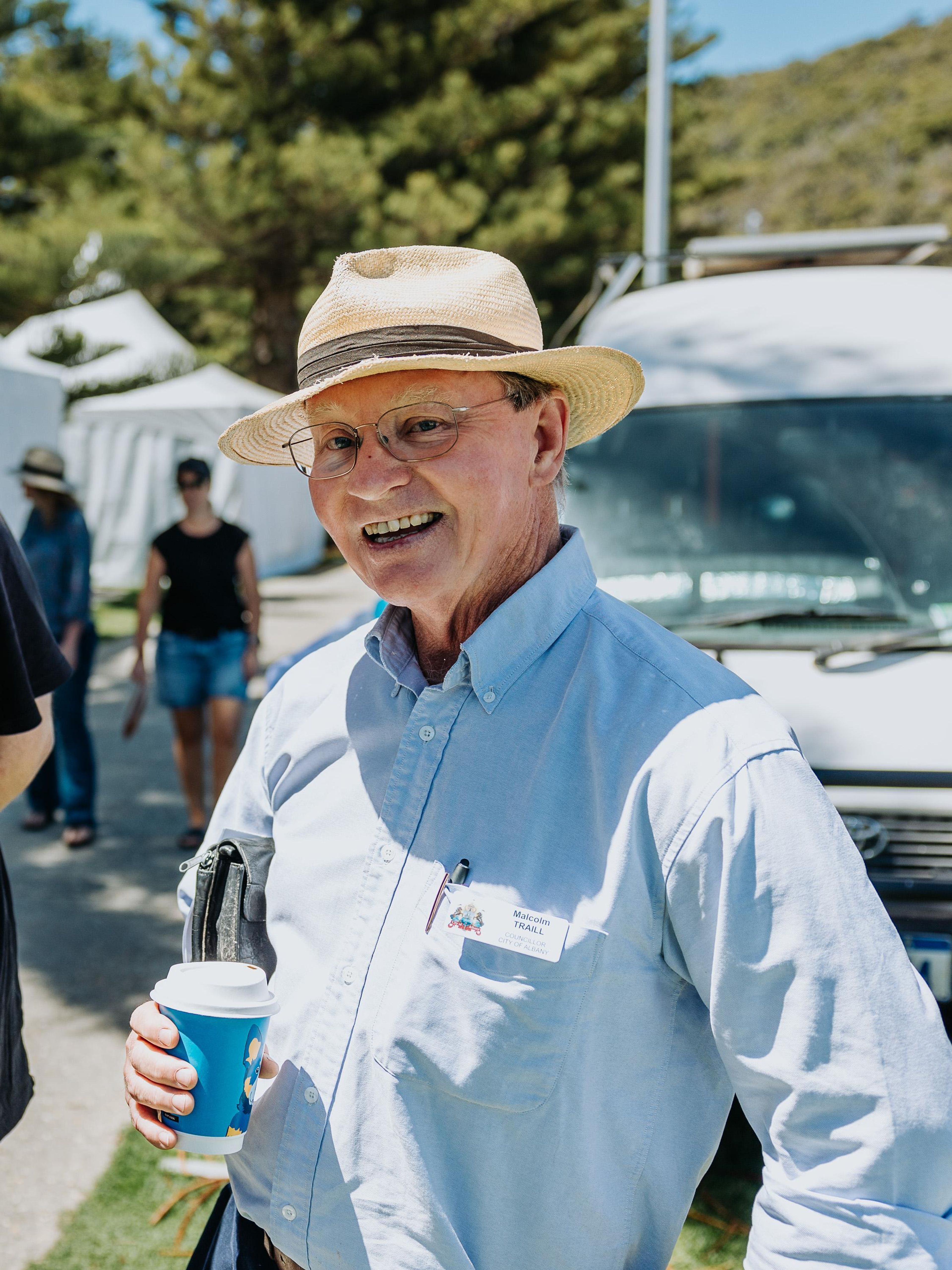 A smiling older man wearing a straw hat, glasses and a light blue shirt holds a takeaway coffee cup while standing outdoors at a sunny event.