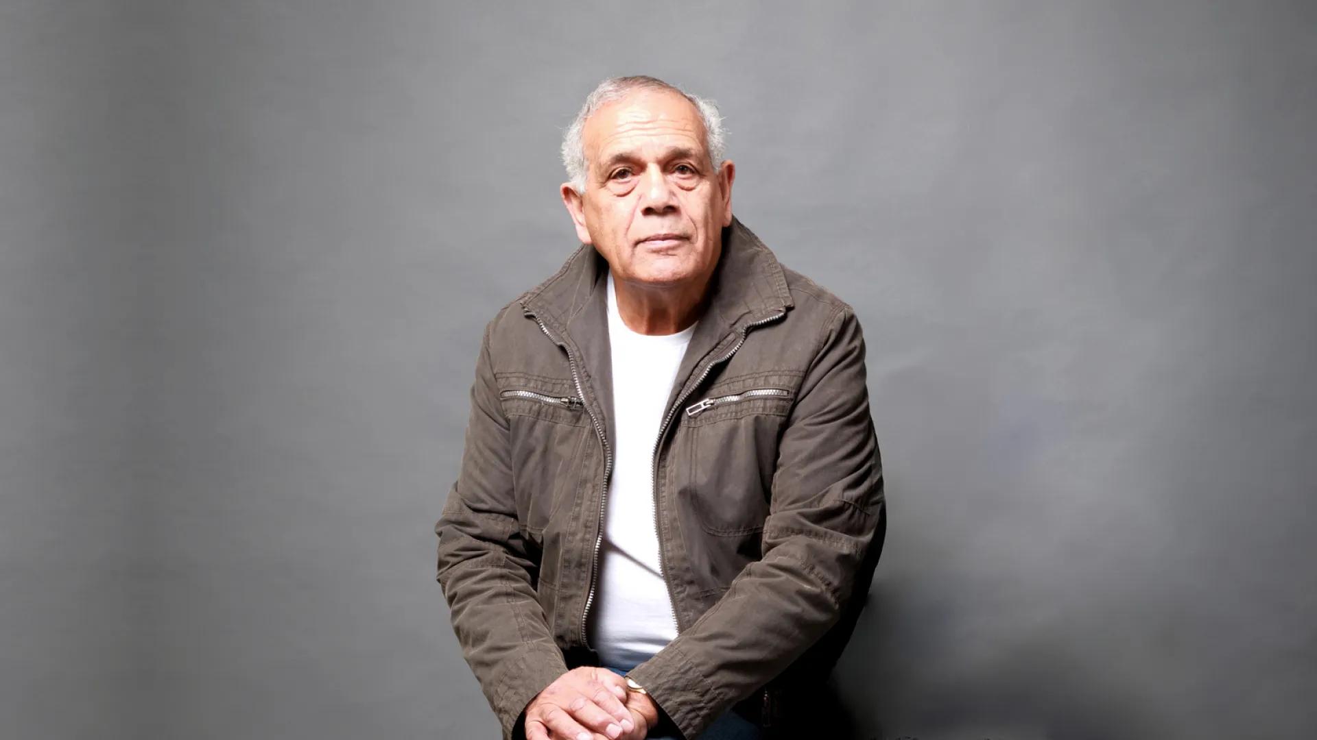 A photo of a man sitting on a chair in front of a grey background.