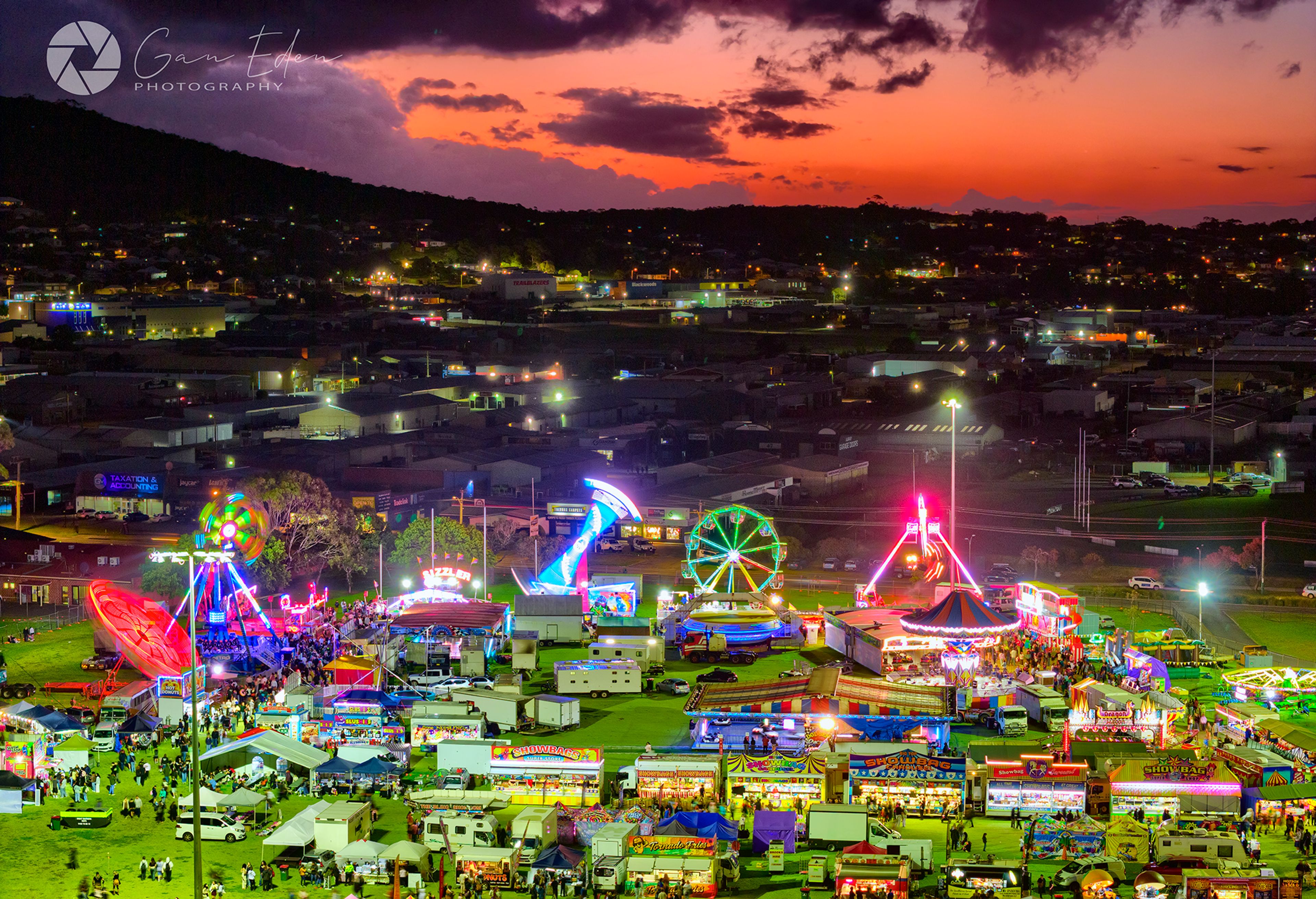 Aerial image of a show ground, with the rides illuminated against a peach-coloured sunset.