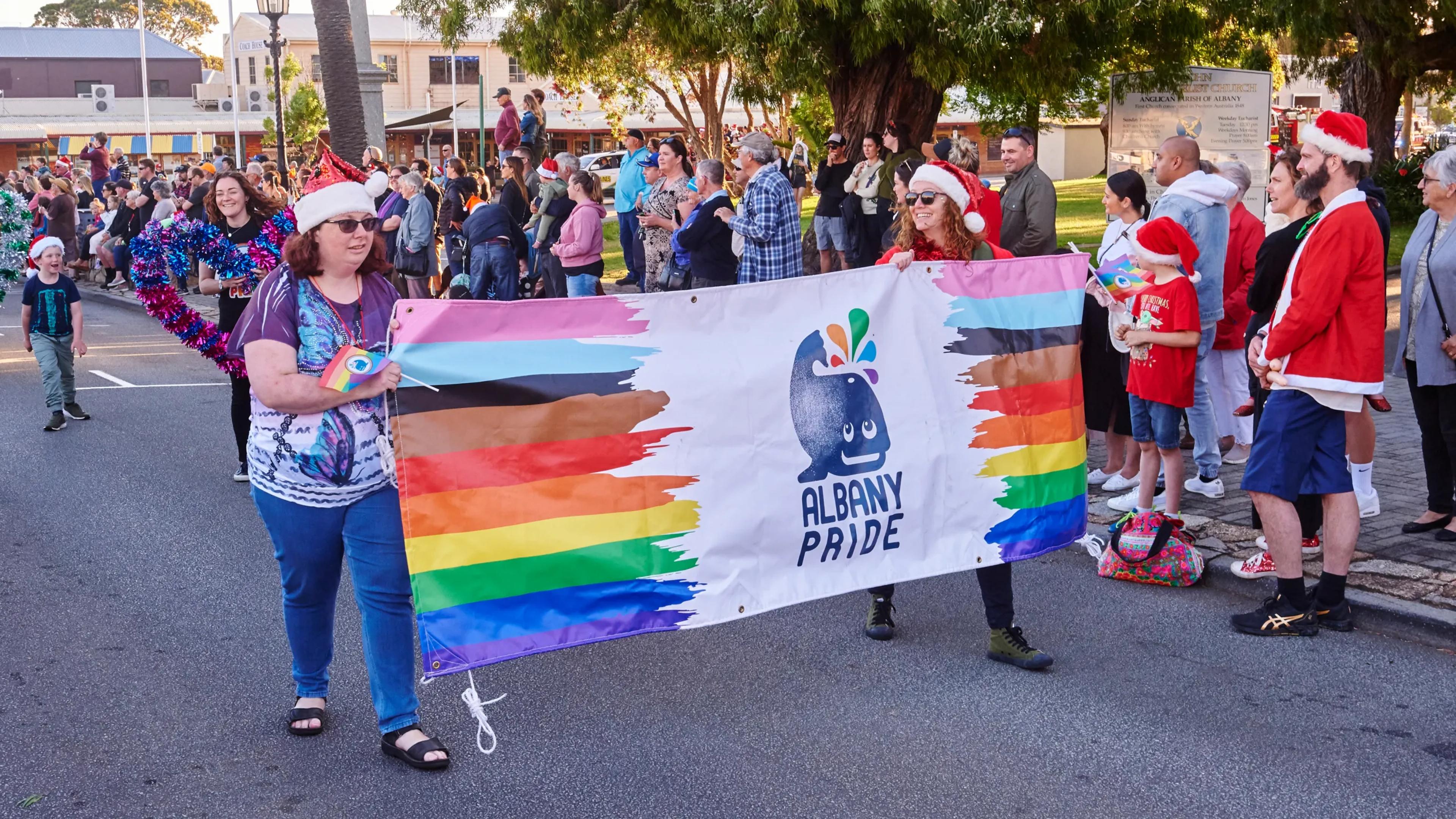 A photo of two women holding an "Albany Pride" flag between them with a series of people behind them standing on the street celebrating.