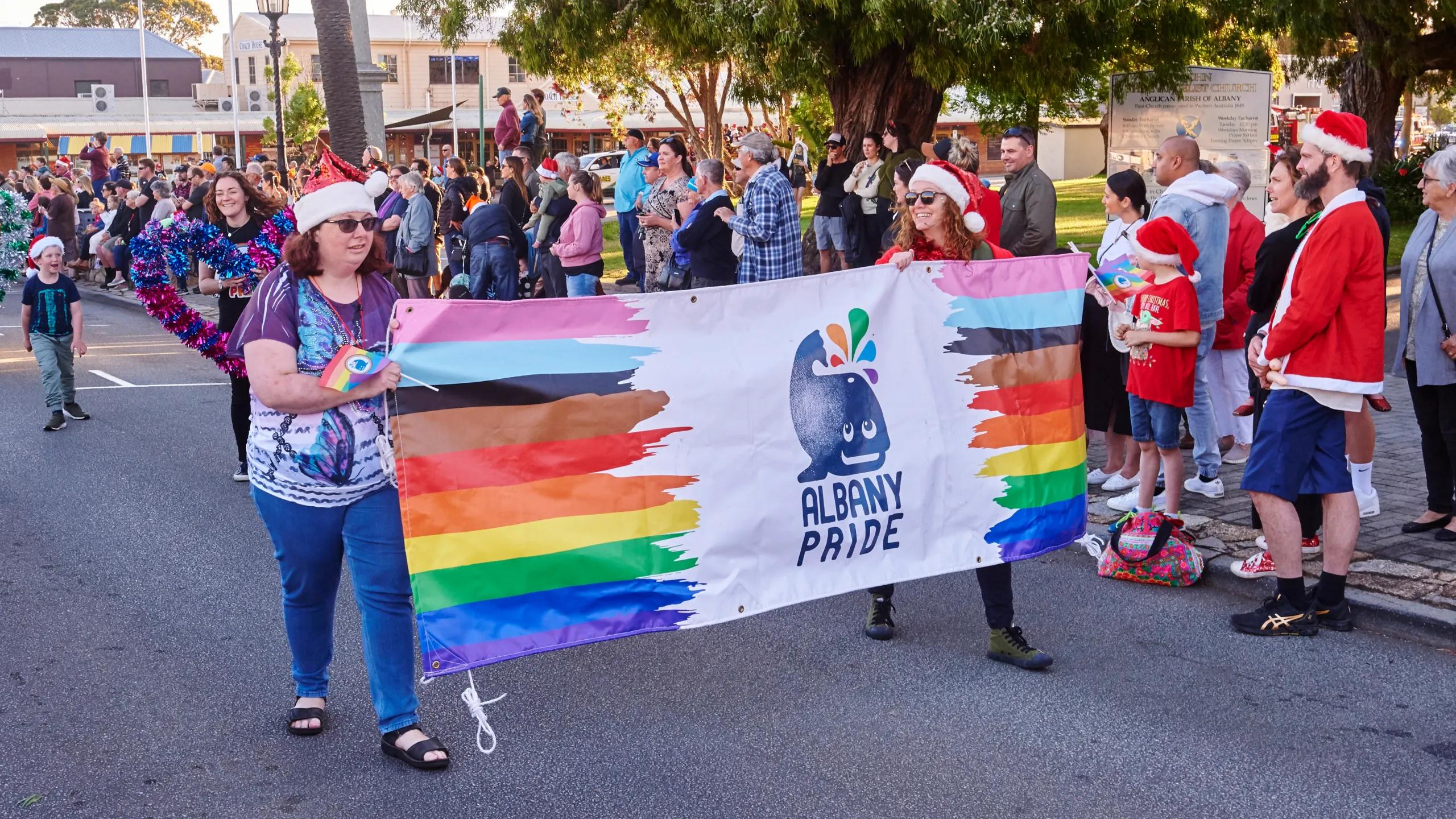 A photo of two women holding an "Albany Pride" flag between them with a series of people behind them standing on the street celebrating.