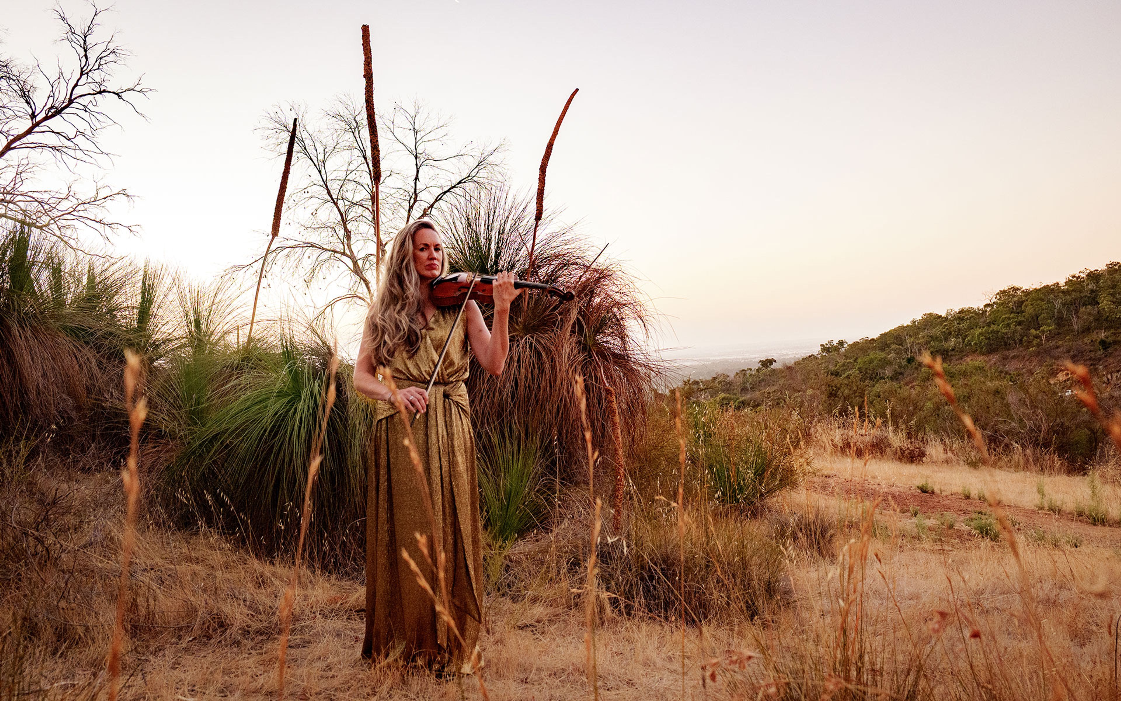 Woman with violin in gold dress stands in Australia bush