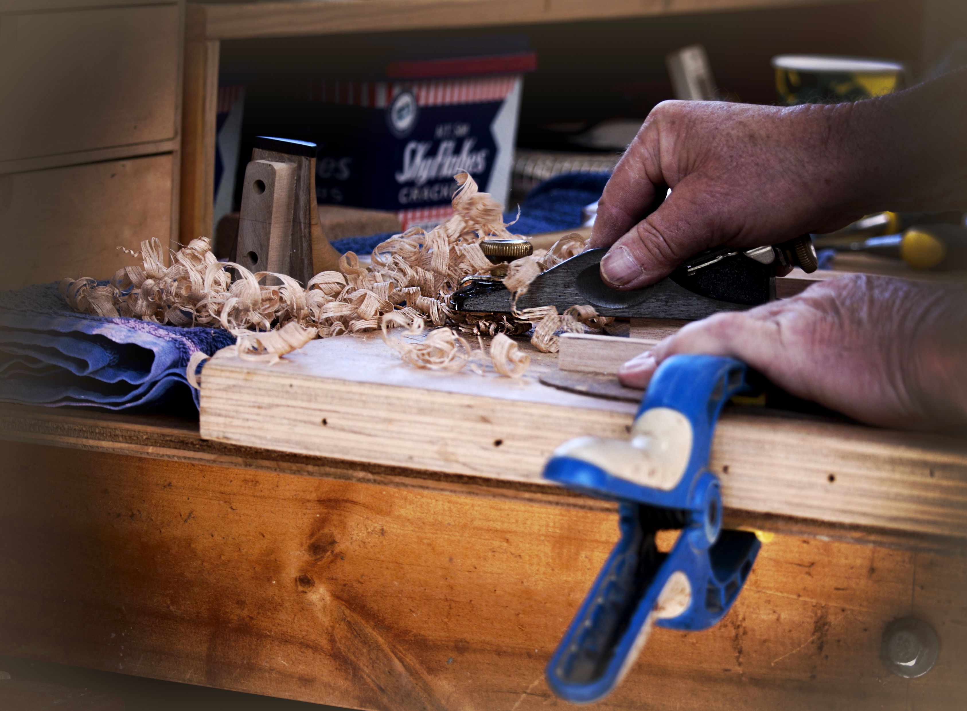 A person’s hands use a small hand plane to shave wood on a workbench, creating curled wood shavings.