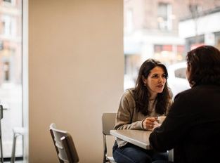 Una donna al tavolino di un bar si gira verso un amico che le sta parlando.