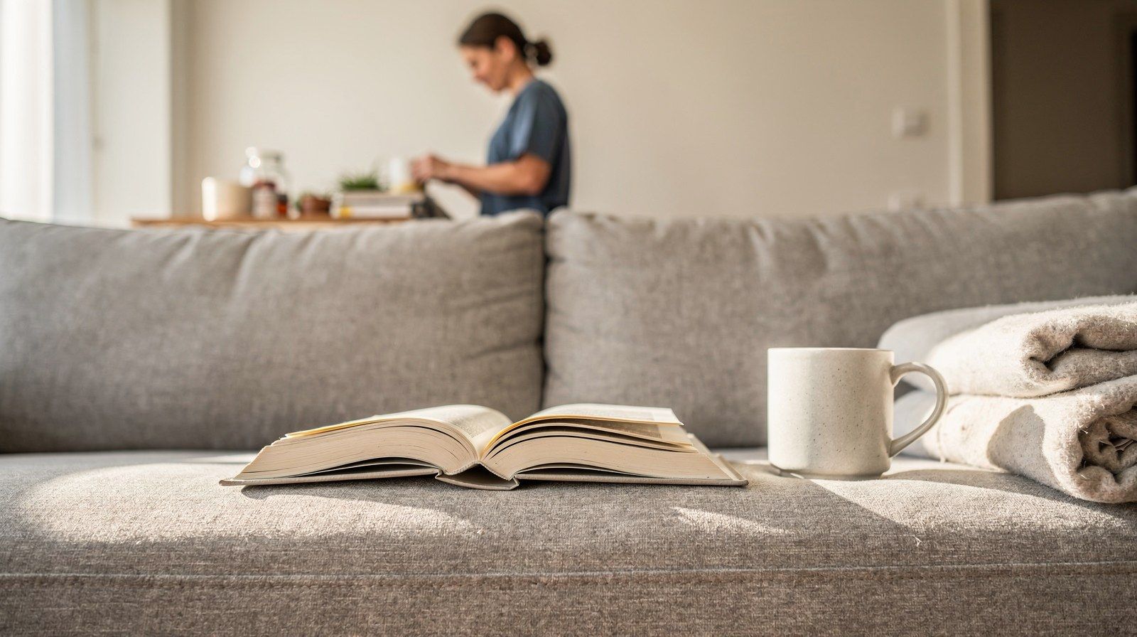 Un libro aperto capovolto su un divano, accanto a una tazza e una coperta piegata.