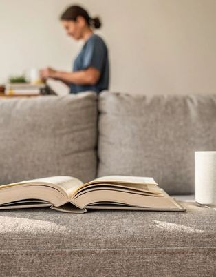 Un libro aperto capovolto su un divano, accanto a una tazza e una coperta piegata.