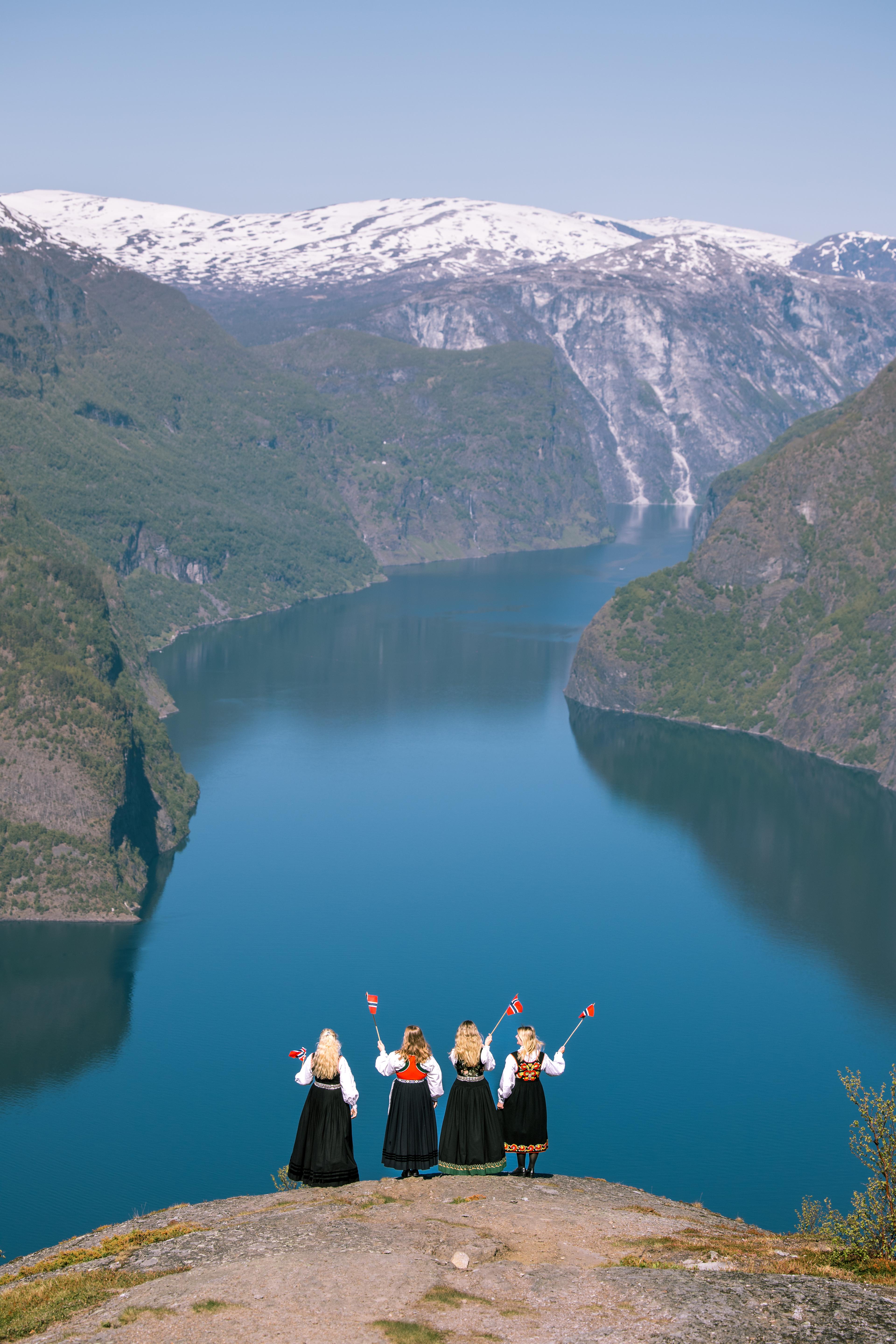 Close-up of a white bunad bodice with delicate folds and purple floral embroidery, with a fjord landscape faintly visible in the background