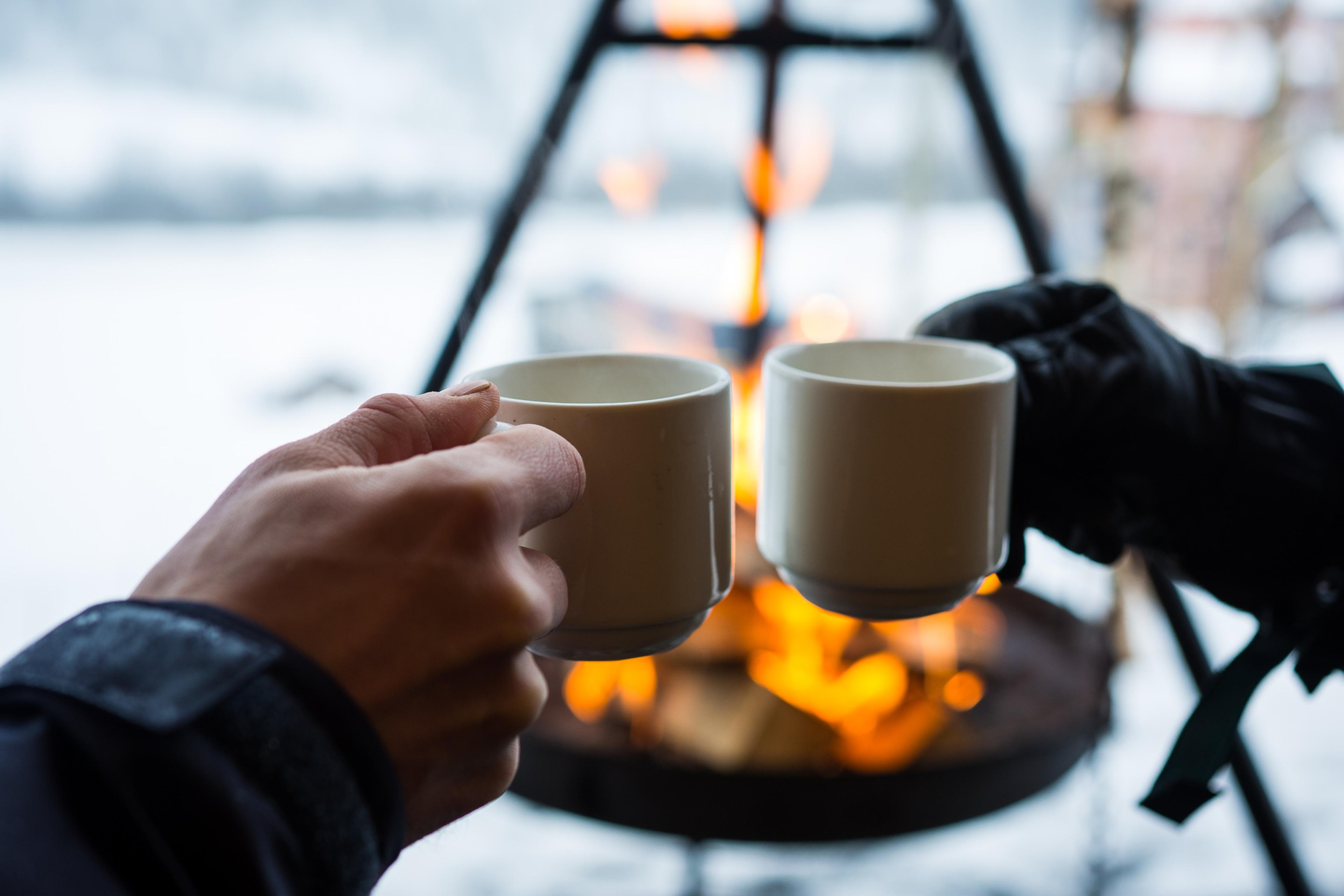 To hender som skåler med kaffekopp foran bålpanna en vinterdag.