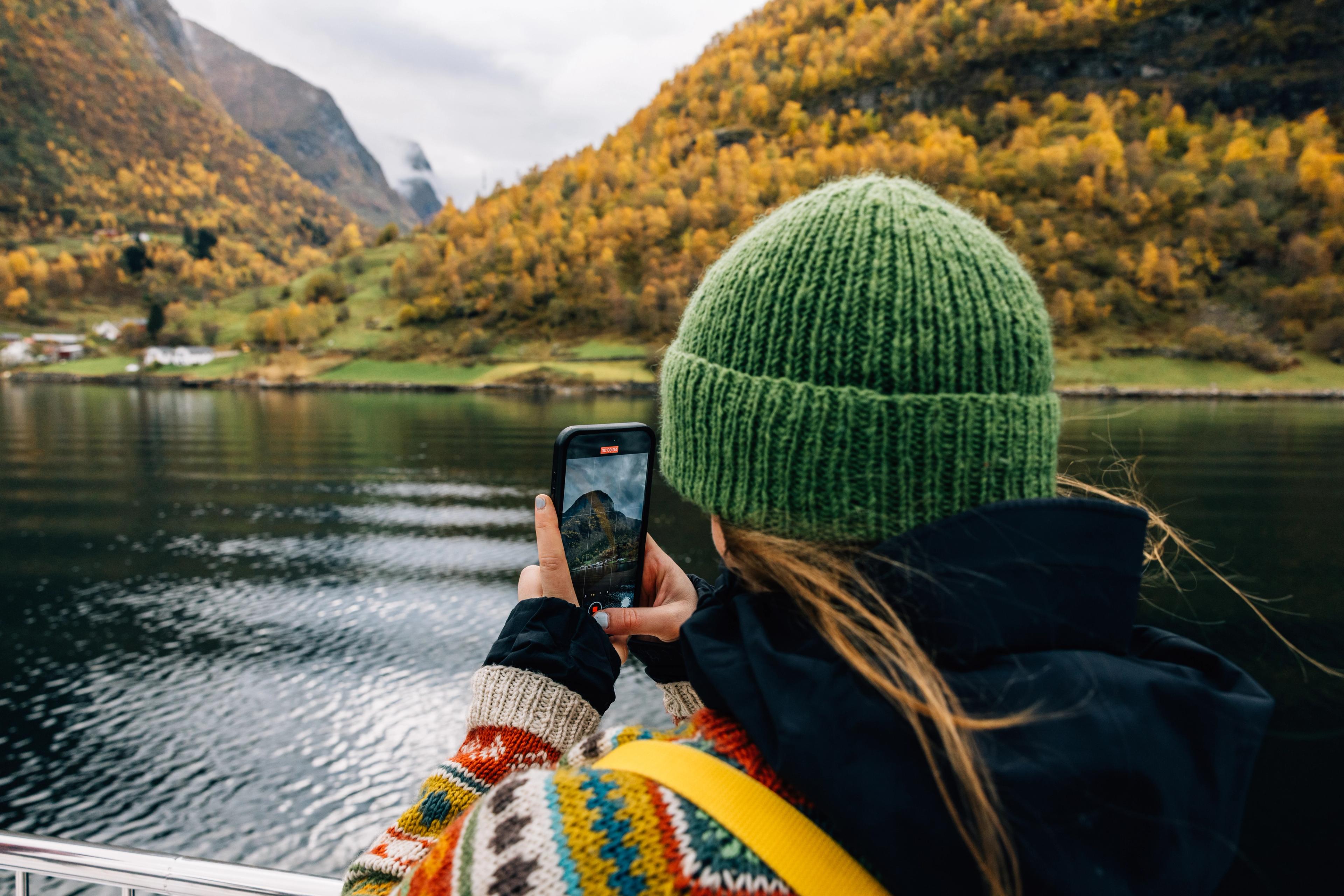 En kvinne i grønn lue og fargerik ullgenser ses bakfra mens hun tar mobilbilde av Undredal fra en båt på Aurlandsfjorden.