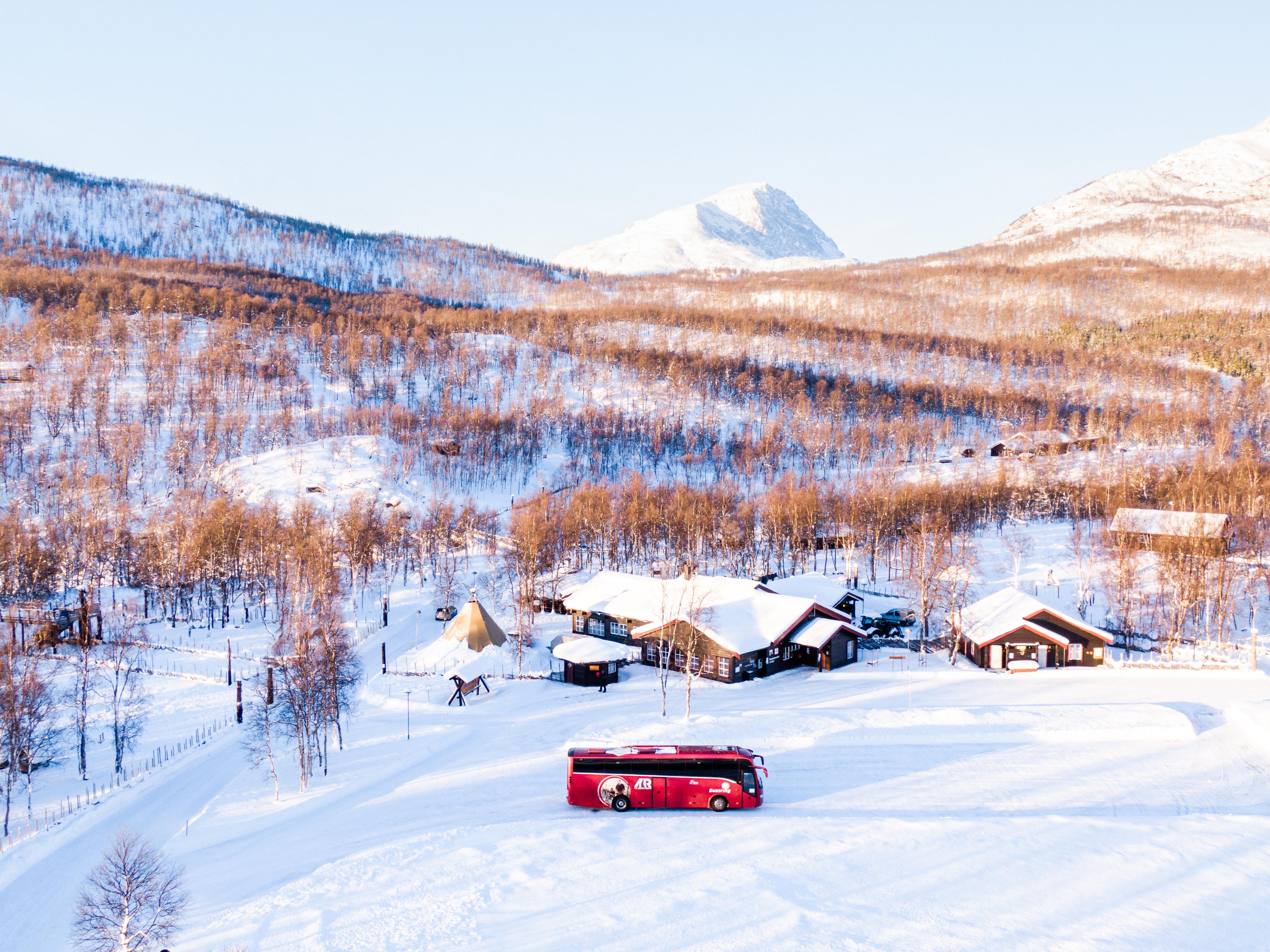 Un bus rouge Arctic Route traverse un paysage enneigé, avec des montagnes et des maisons à l’arrière-plan.