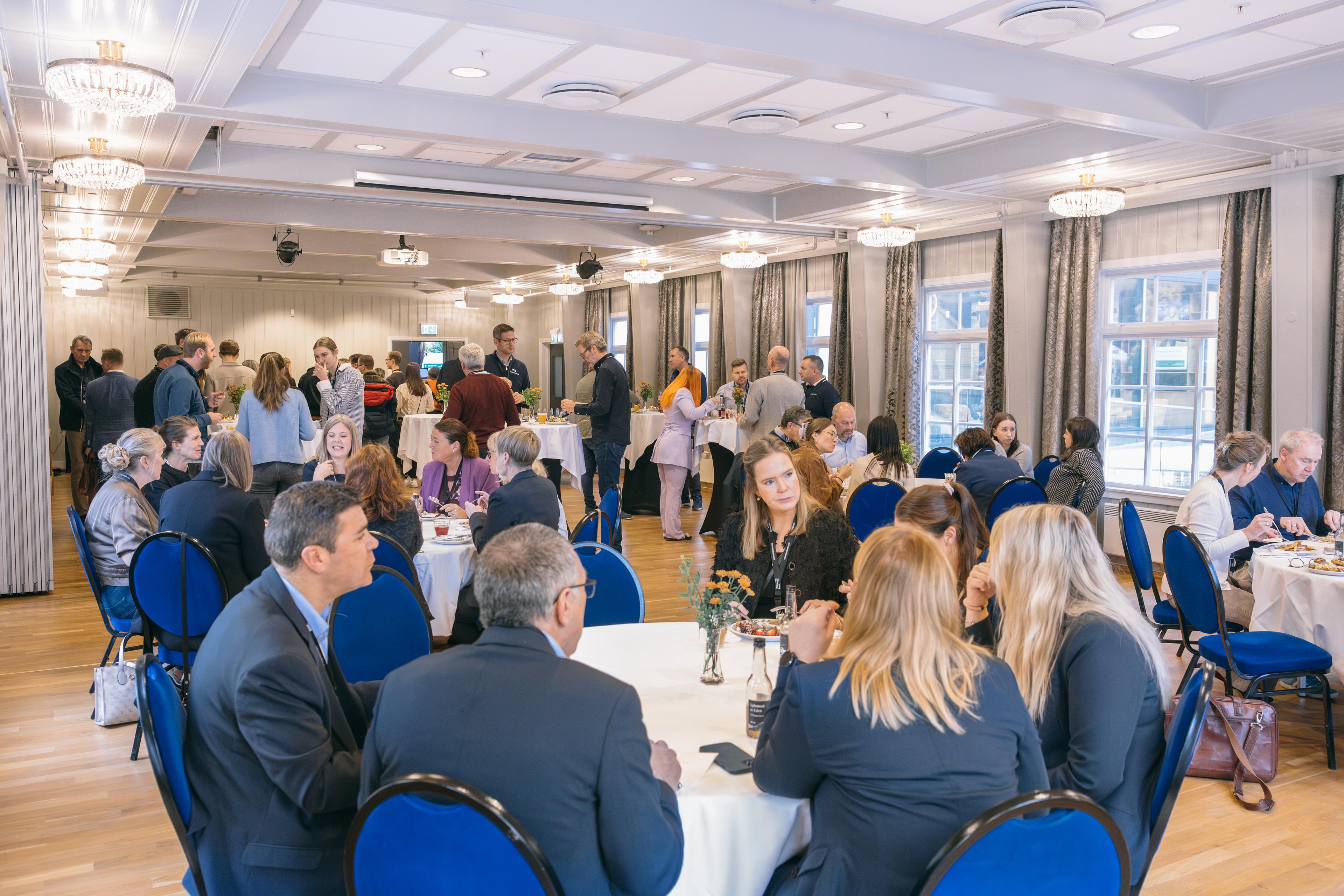 Deltakarar i samtale og mingling under Næringskonferansen, med bordservering og ståbord i ein lys konferansesal.
