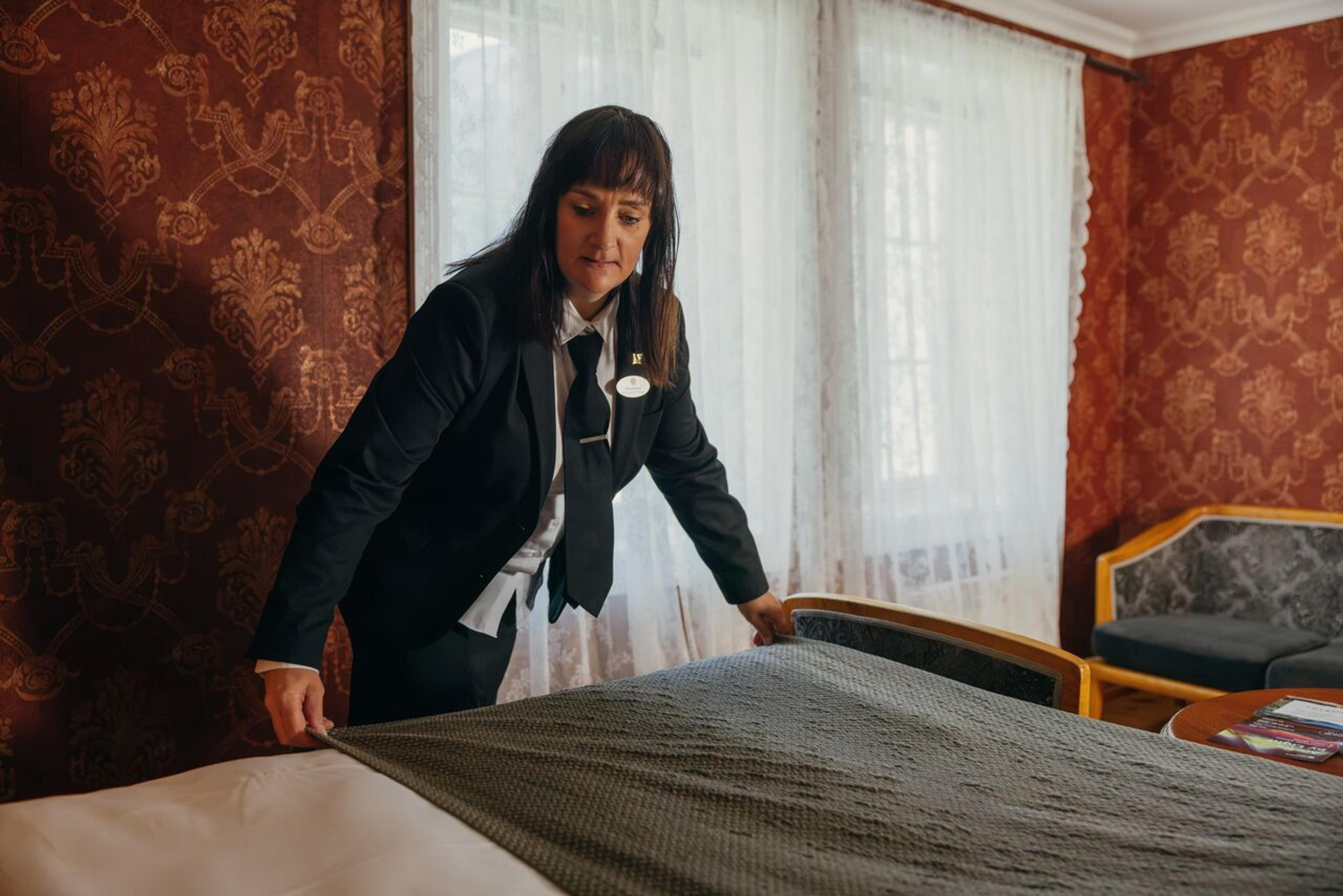 A woman in a hotel uniform adjusts a bedspread on a hotel bed.
