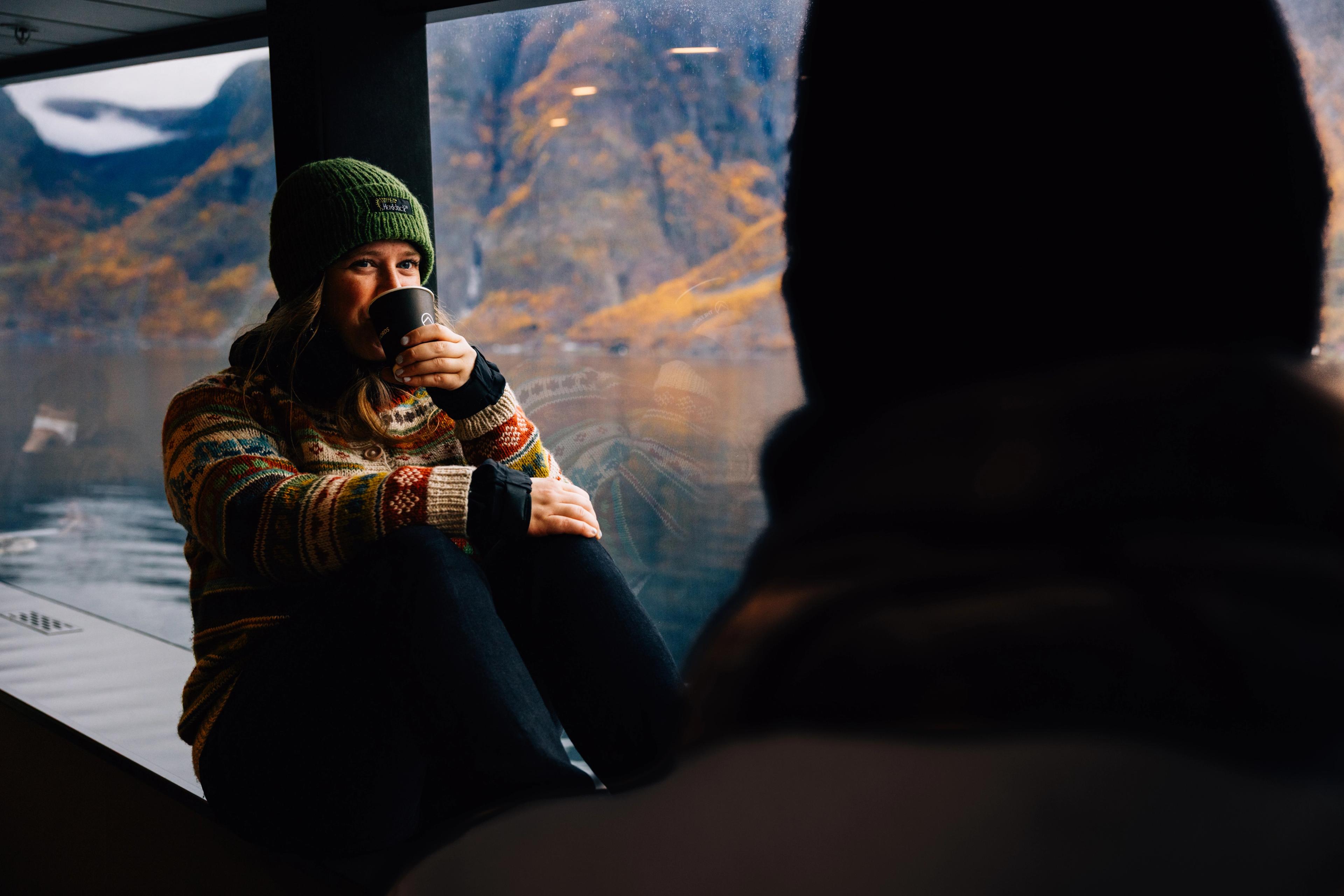En kvinne sitter i vinduskarmen om bord på en båt og tar en slurk kaffe mens hun smiler lurt mot en annen passasjer man bare ser bakhodet til. Høstfargete fjellsider utenfor.