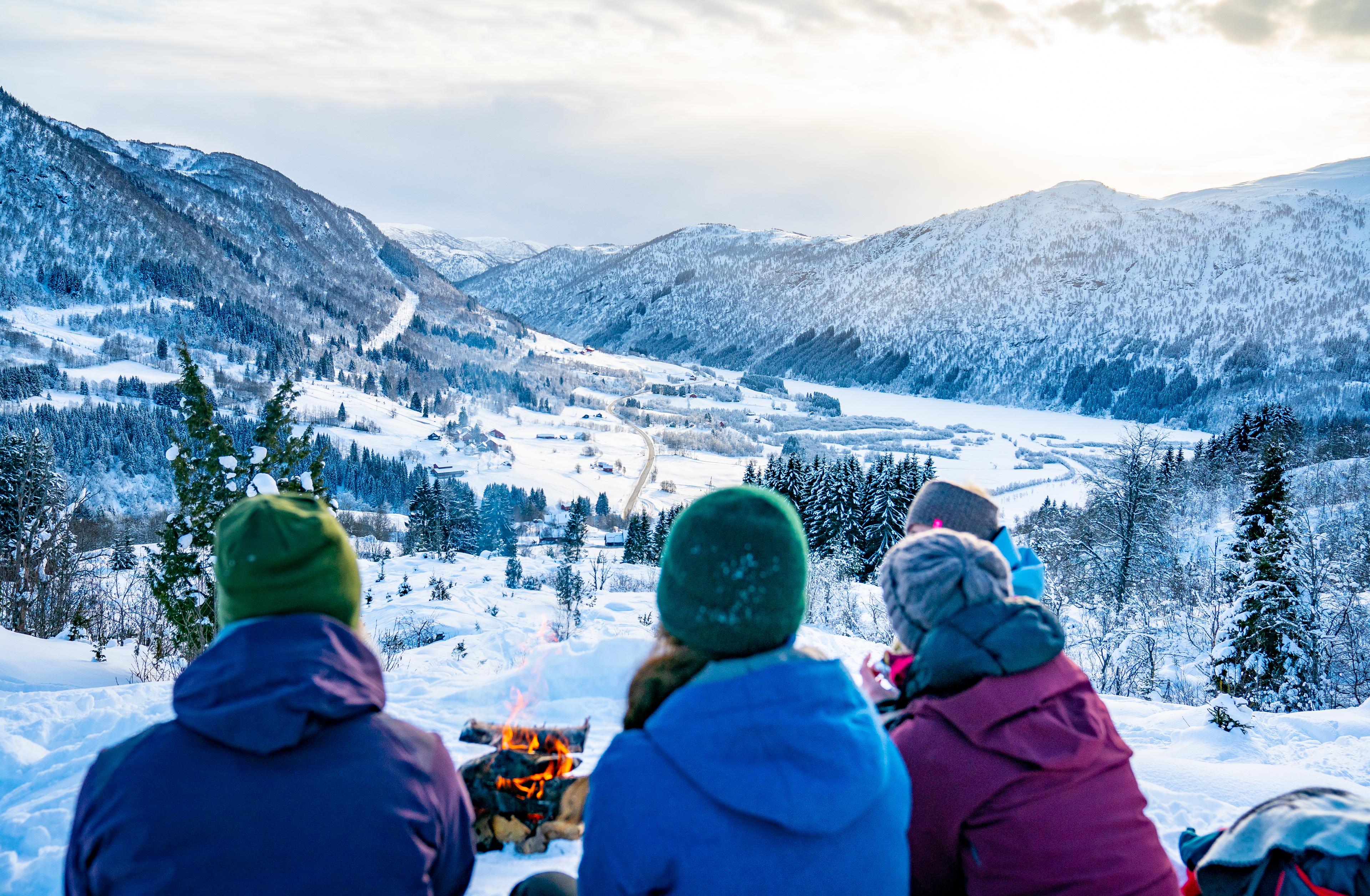 Friends on a trip sit by a fire and have an adventurous view of the snow-covered valley.