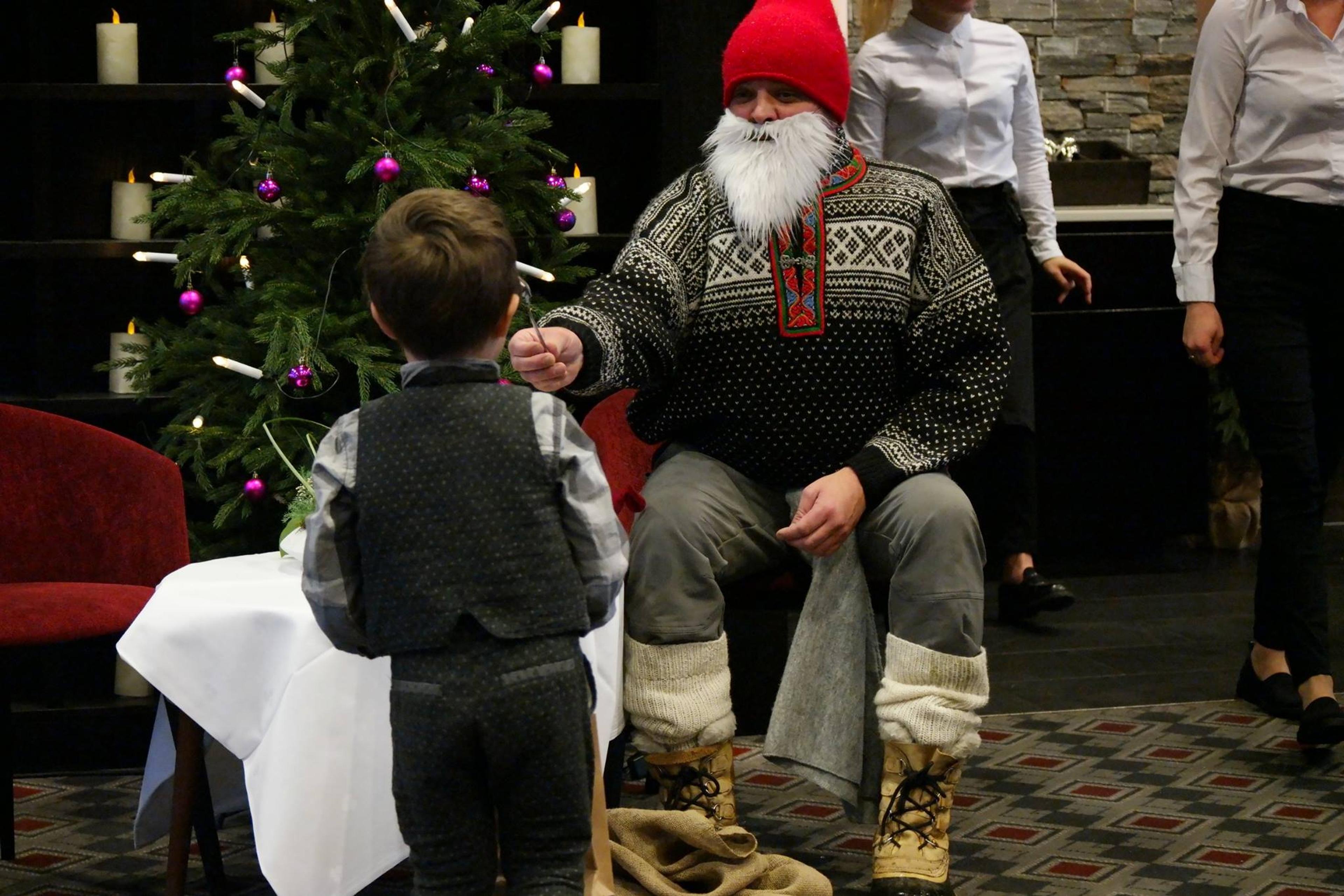 En julenisse rekker noe til en gutt som står med ryggen til på bilde. Nissen sitter ved et juletre.
