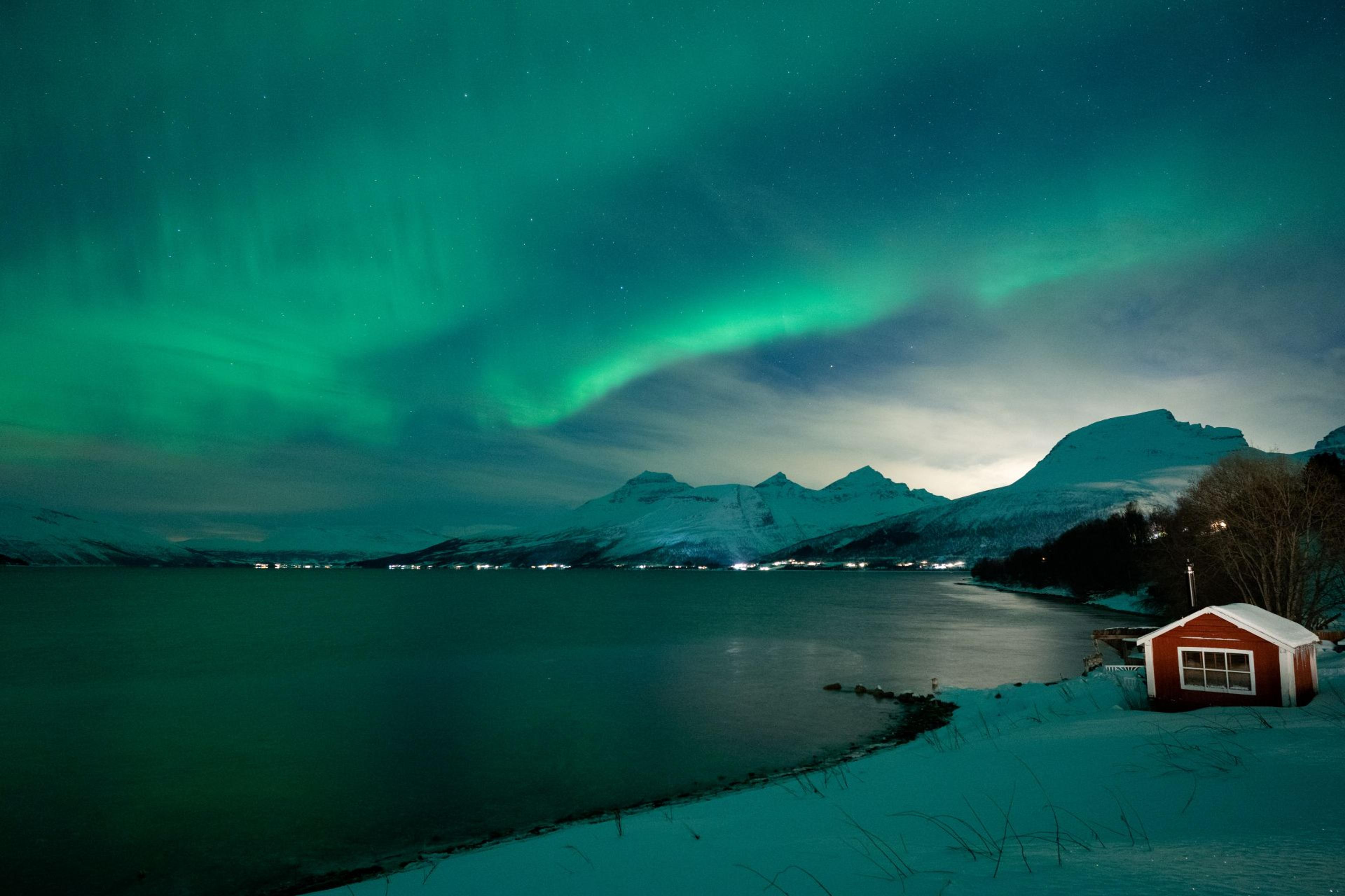 Northern Lights over the fjord and snow-covered mountains, with a red boathouse in the foreground.