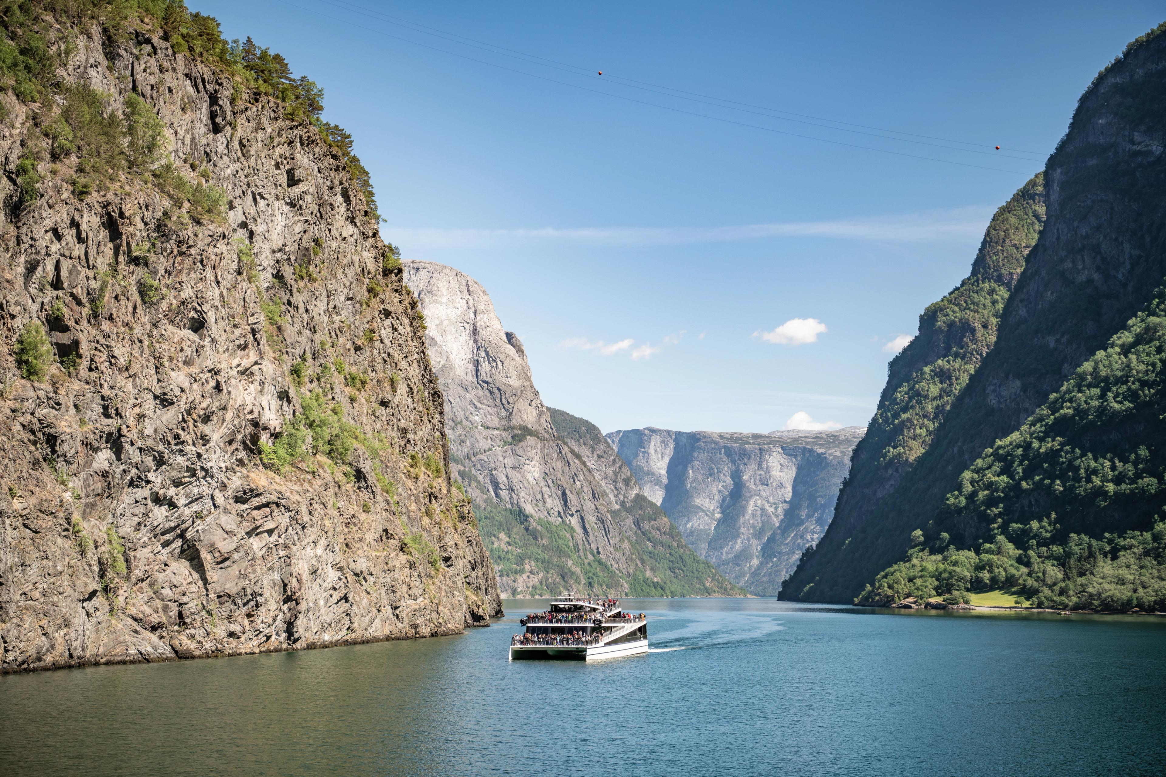 En sightseeingbåt på fjorden omringet av bratte fjell og mange gjester på dekk. Himmelen viser en klar sommerdag.