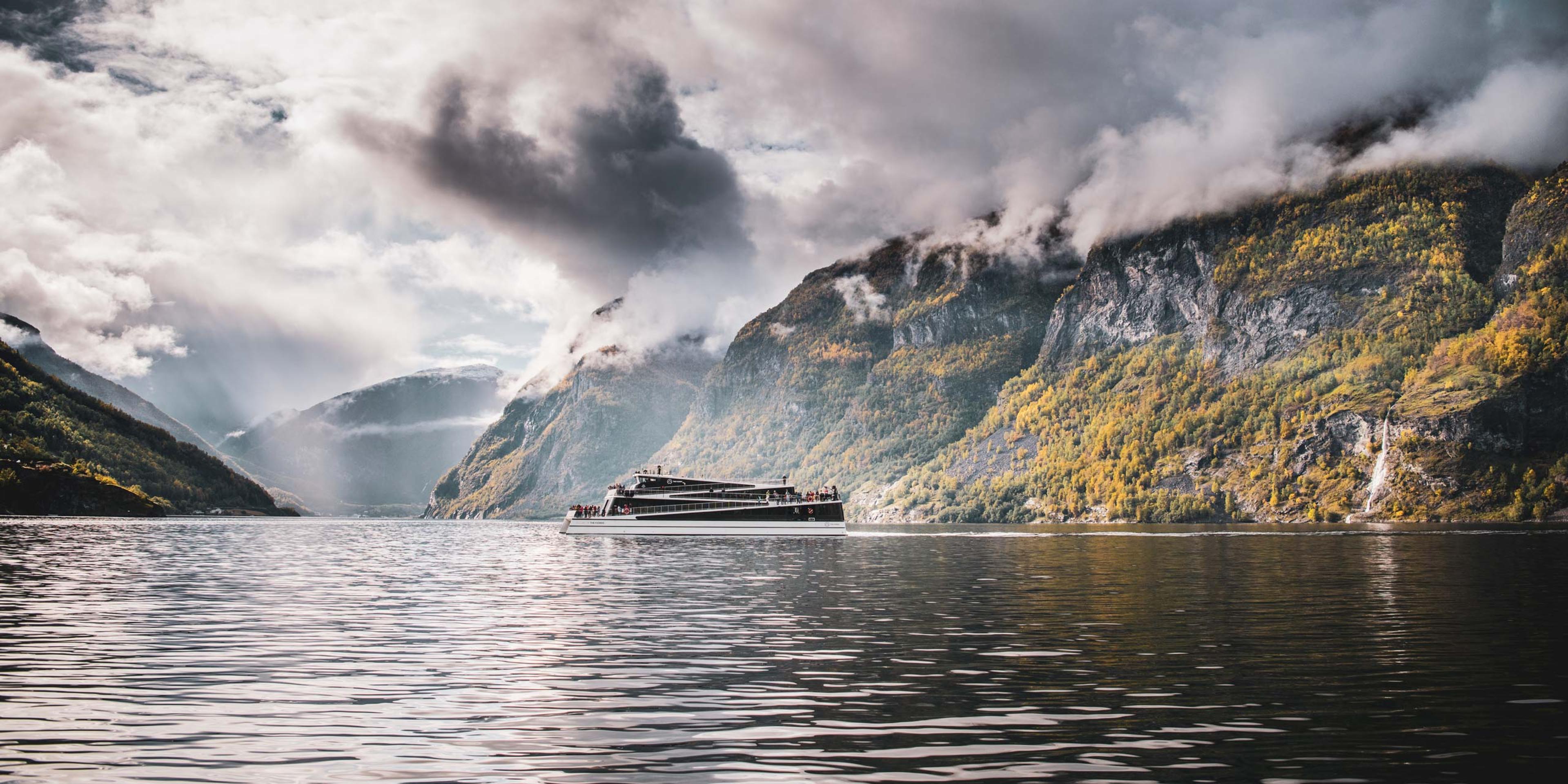 A fully electric cruise ship sailing through the calm waters of Nærøyfjord in Norway, surrounded by steep mountain cliffs. The ship operates silently, showcasing its eco-friendly, battery-powered design to minimize environmental impact on the UNESCO-listed fjord.