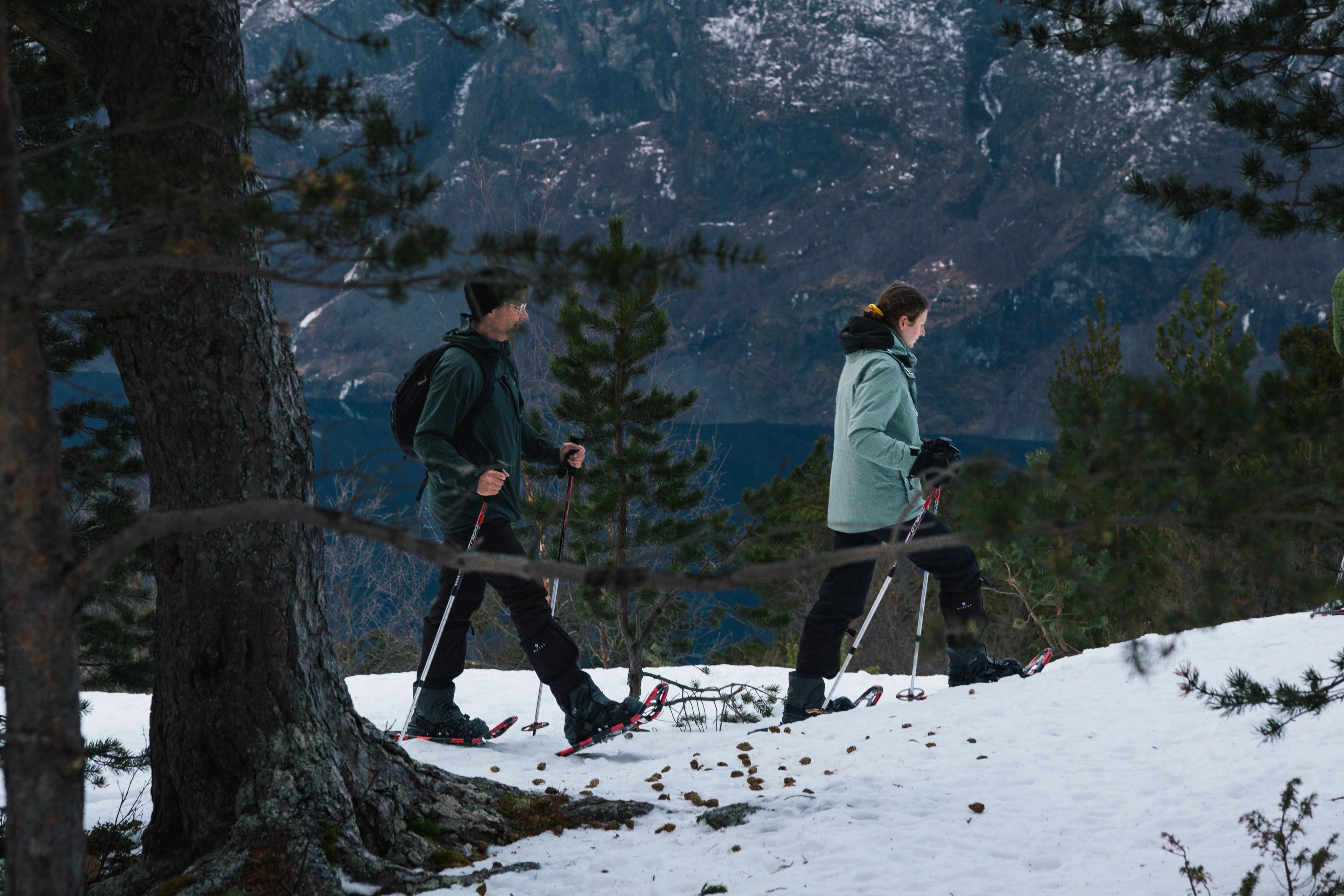  A man and a woman are snowshoeing in open forest with a view of the Aurlandsfjord behind.