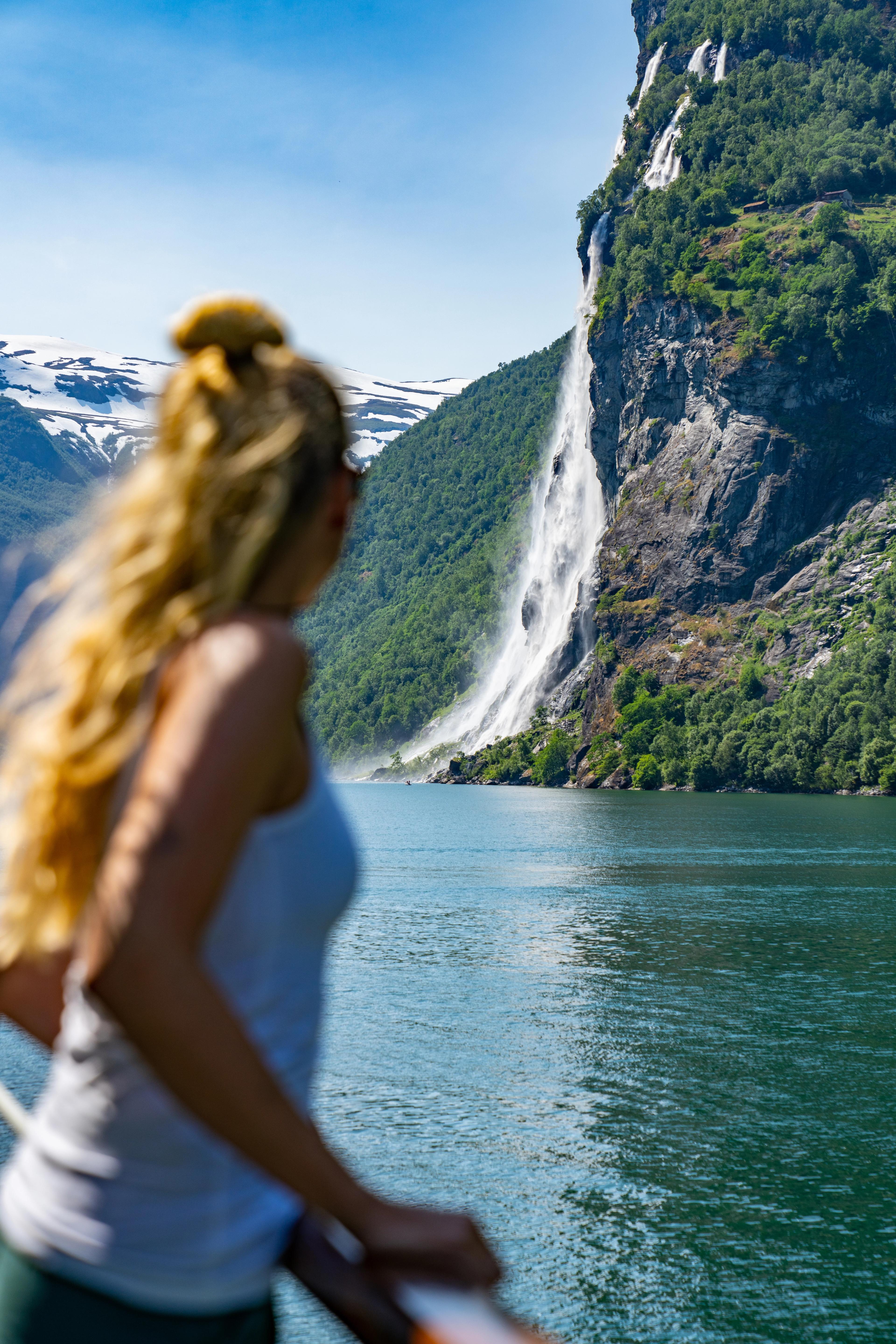 A woman on a boat looking at a waterfall in summer. The fjord is turquoise, with snow-covered mountains in the background.