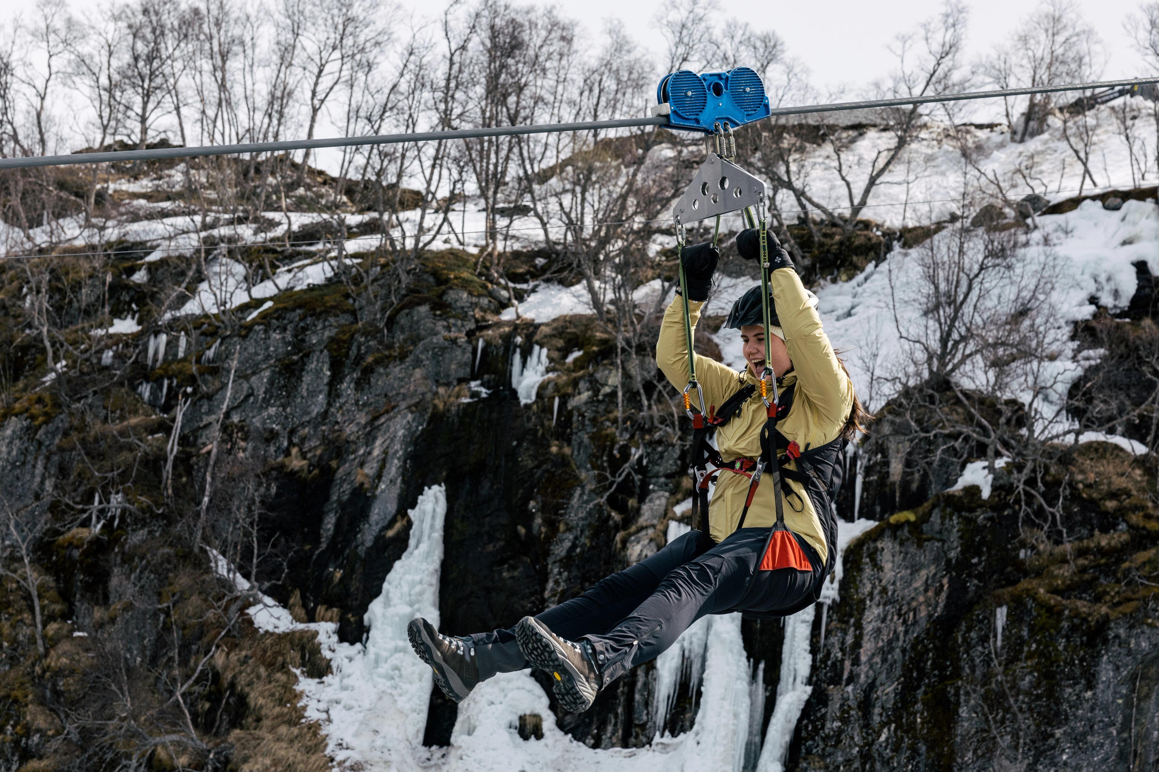 Une femme suspendue dans les airs sur la tyrolienne de Flåm au début du printemps, avec de la glace et de la neige visibles sur les falaises en arrière-plan.