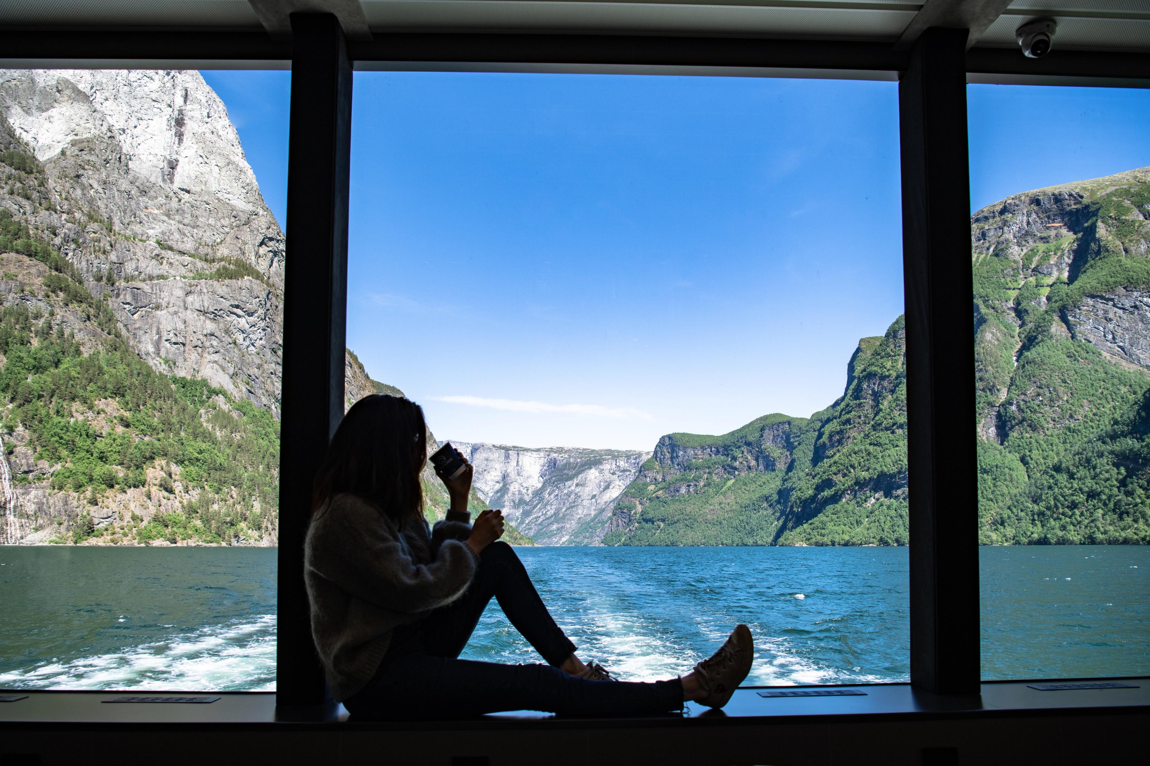 Silhuetten av en kvinne som sitter i et panoramavidu mot fjorden. På vannet ser man bølgene etter båten ho sitter i.